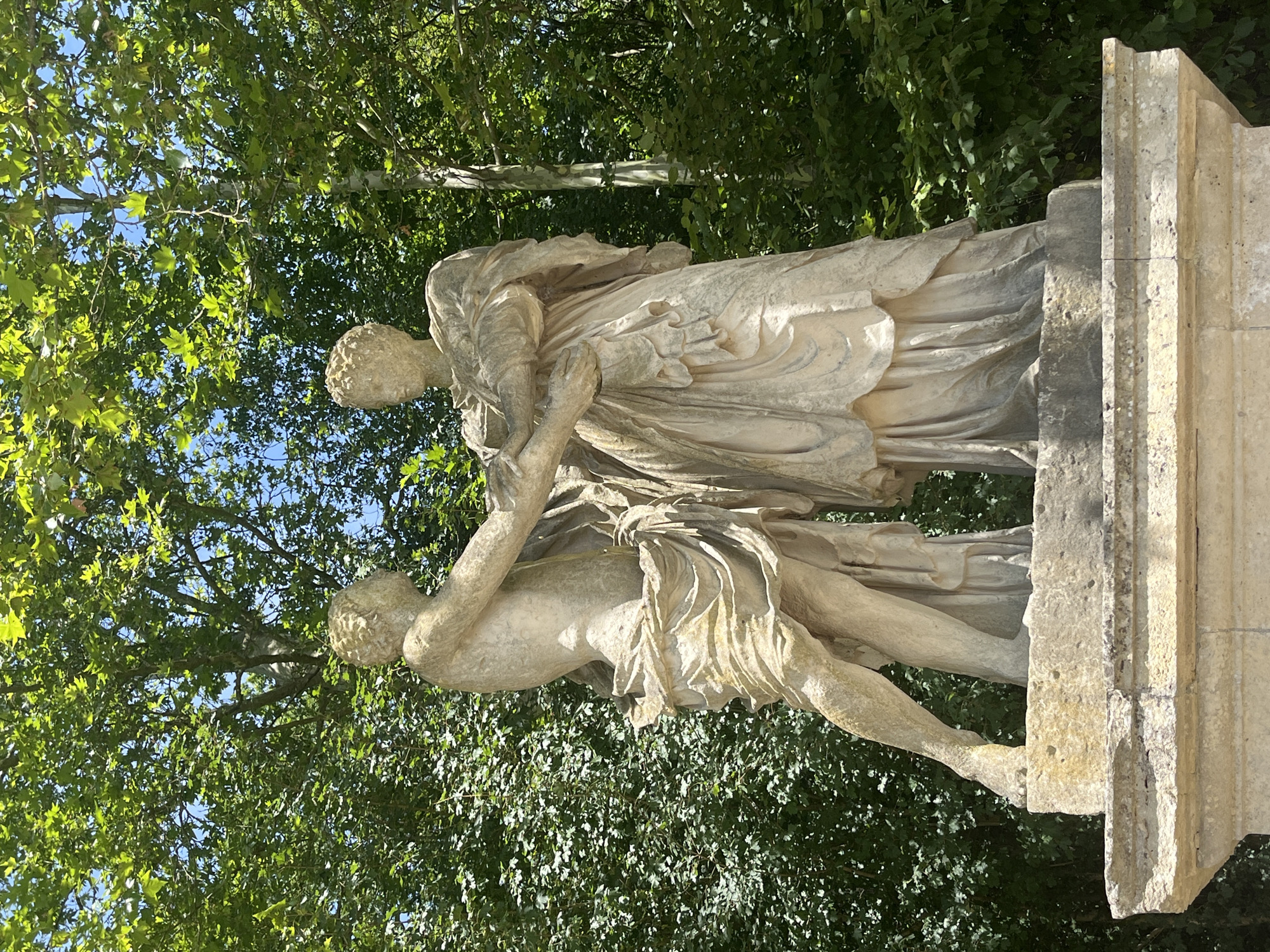 This image depicts a marble sculpture of two figures in the Parc de Sceaux, located in Antony, France. The sculpture is positioned on a stone pedestal amidst lush green foliage and a clear blue sky. The figures appear to be in an embrace, with one figure, presumably male, reaching out to the other, presumably female. Both figures are draped in flowing classical attire, rendered with intricate detail in the marble. The sculpture shows signs of age and weathering, characteristic of outdoor art. The vibrant green leaves of the surrounding trees create a natural backdrop, contrasting with the pale stone of the artwork. The sunlight casts subtle shadows, highlighting the textures and contours of the sculpture.