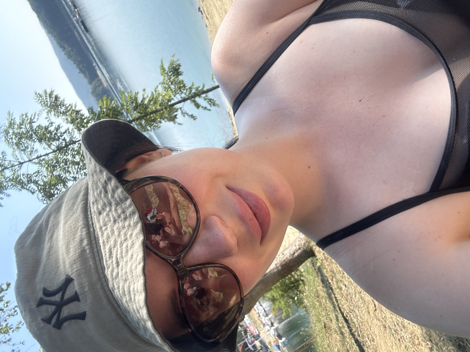 In Polańczyk, Poland, a woman is captured in a selfie by a lake. She is wearing a light-colored bucket hat with a New York Yankees logo and dark sunglasses. The sunglasses reflect a scene of a person sitting with flowers in their hair and a colorful patterned fabric. She is also wearing a black bikini top. The background shows a tranquil lake with distant hills and boats docked along the shore, suggesting a leisurely outdoor setting. The sun is shining, casting a warm glow on the scene.
