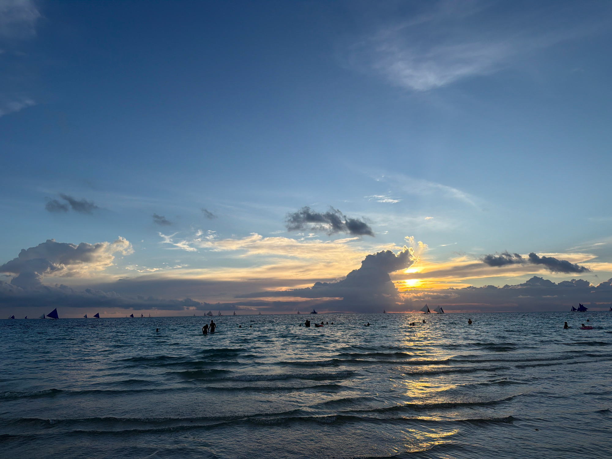 An ocean sunset scene at Boracay Beach in Malay, Philippines shows a vibrant sky with hues of orange, yellow, and blue, painted with scattered clouds. The calm sea reflects the sky's colors, with gentle waves lapping at the shore. Several sailboats, with triangular purple sails, are scattered across the water. A few people are visible in the water, some swimming and others standing. The overall atmosphere is serene and picturesque.