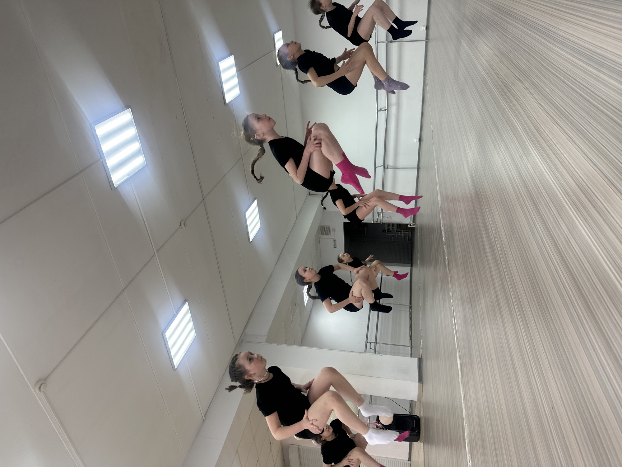 The image shows a dance studio in Votkinsk, Russia, with four girls wearing black clothing and variously colored socks practicing a dance routine. The studio has a mirrored wall reflecting the girls and the interior, including the ceiling with fluorescent lights. The floor appears to be covered in light-colored striped wallpaper, and there is a ballet barre visible along the mirrored wall.