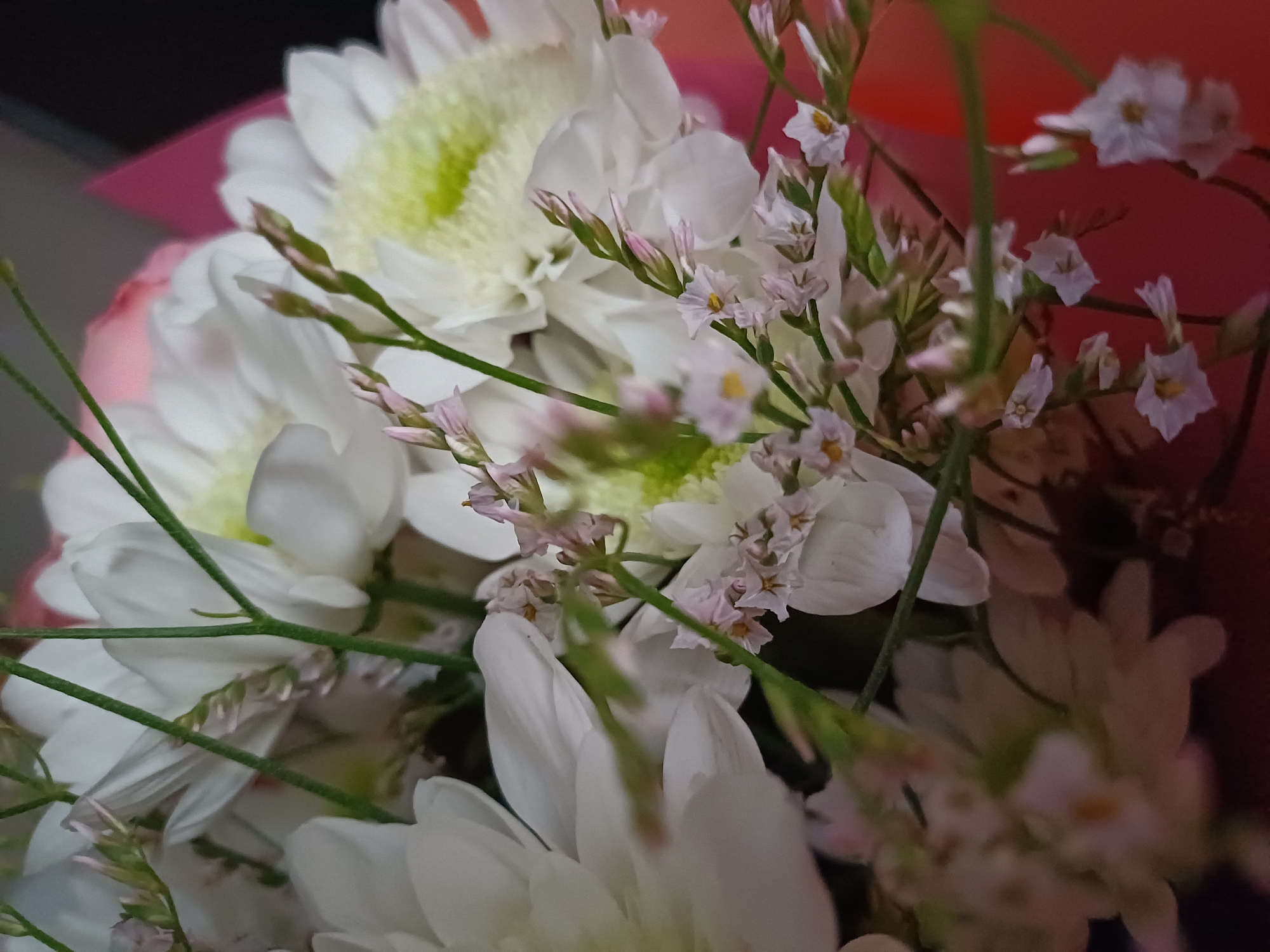 This is a close-up, overhead, vertical shot of a bouquet of white flowers. The flowers are layered and overlapping, with some appearing fully bloomed and others still in bud form. Delicate green stems and small, pale pink buds are interspersed throughout the bouquet. The background is blurred, with a hint of pink wrapping paper visible on the right side of the frame. The lighting is soft and diffused.