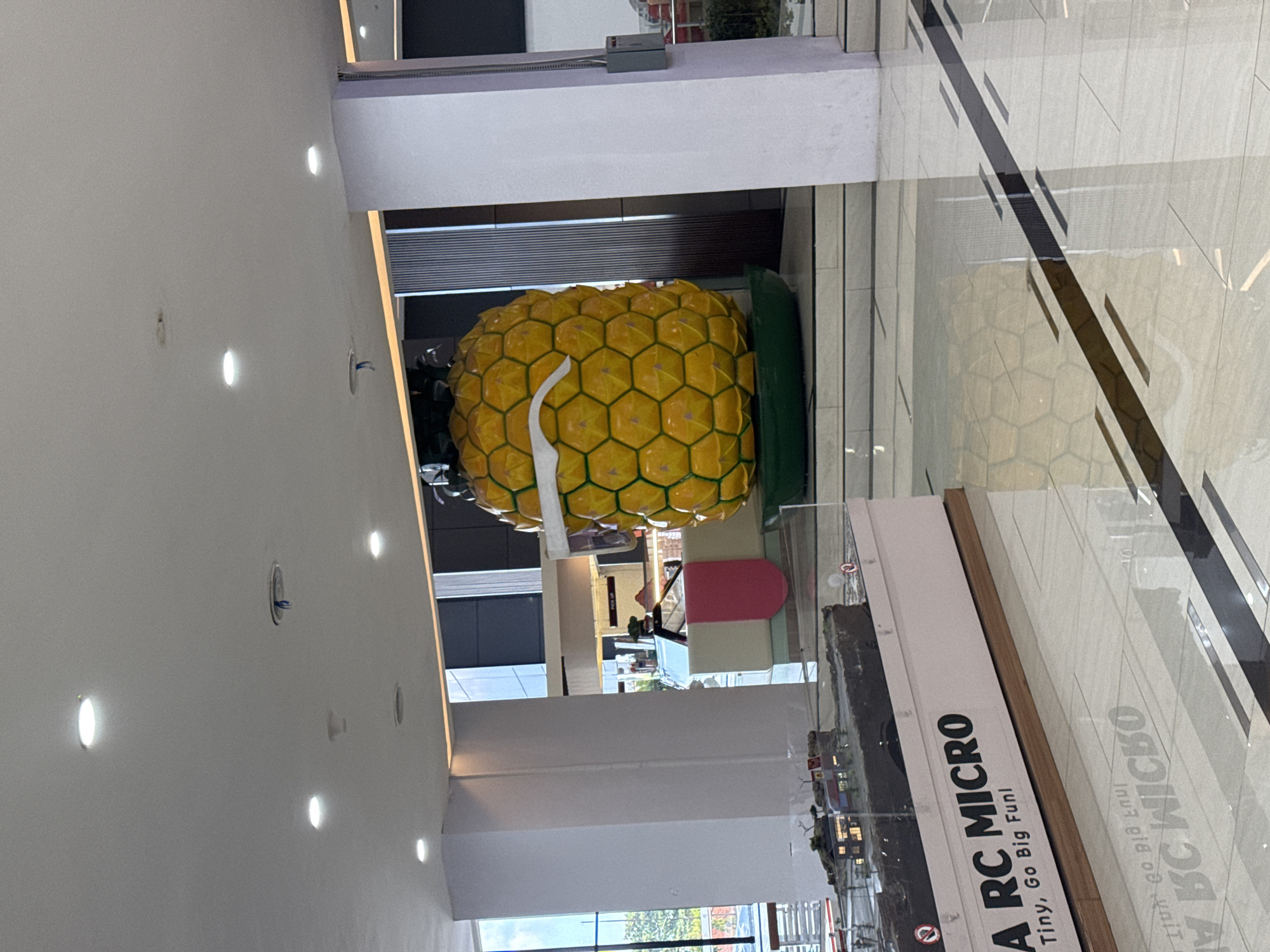 The image shows a large, stylized pineapple sculpture with sunglasses on display indoors, possibly in a mall or public building. The sculpture is yellow with hexagonal segments and sits on a green base. In the foreground, there is a display case with miniature models, one of which is labeled "A RC MICRO Tiny, Go Big Fun!". The floor is tiled and reflects the surrounding environment. The background shows a storefront or service counter with people potentially working behind it. The architecture includes large white columns and a modern ceiling with recessed lighting. The overall scene suggests a commercial or entertainment venue. The presence of the RC Micro display and the large pineapple sculpture hints at a location catering to families or hobbyists. The image was taken at the Discovery Mall in Kuta, Indonesia.