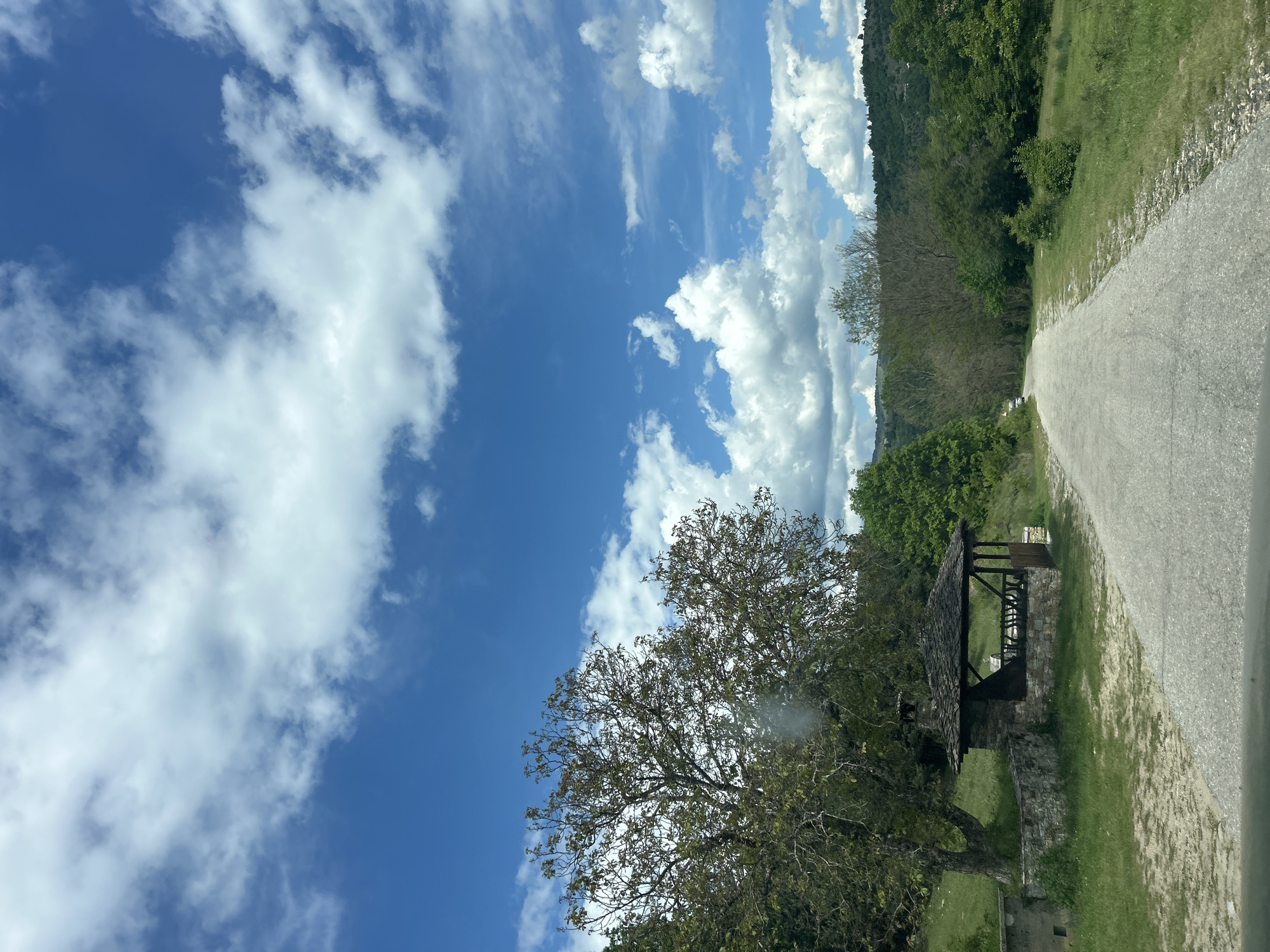 The image shows a section of a road next to an area with trees, a small stone structure, and a cloudy sky. The road is paved and appears to be a light gray color. The structure is built from stone and wood. The sky is blue with white clouds. The location is Pletena, Bulgaria.