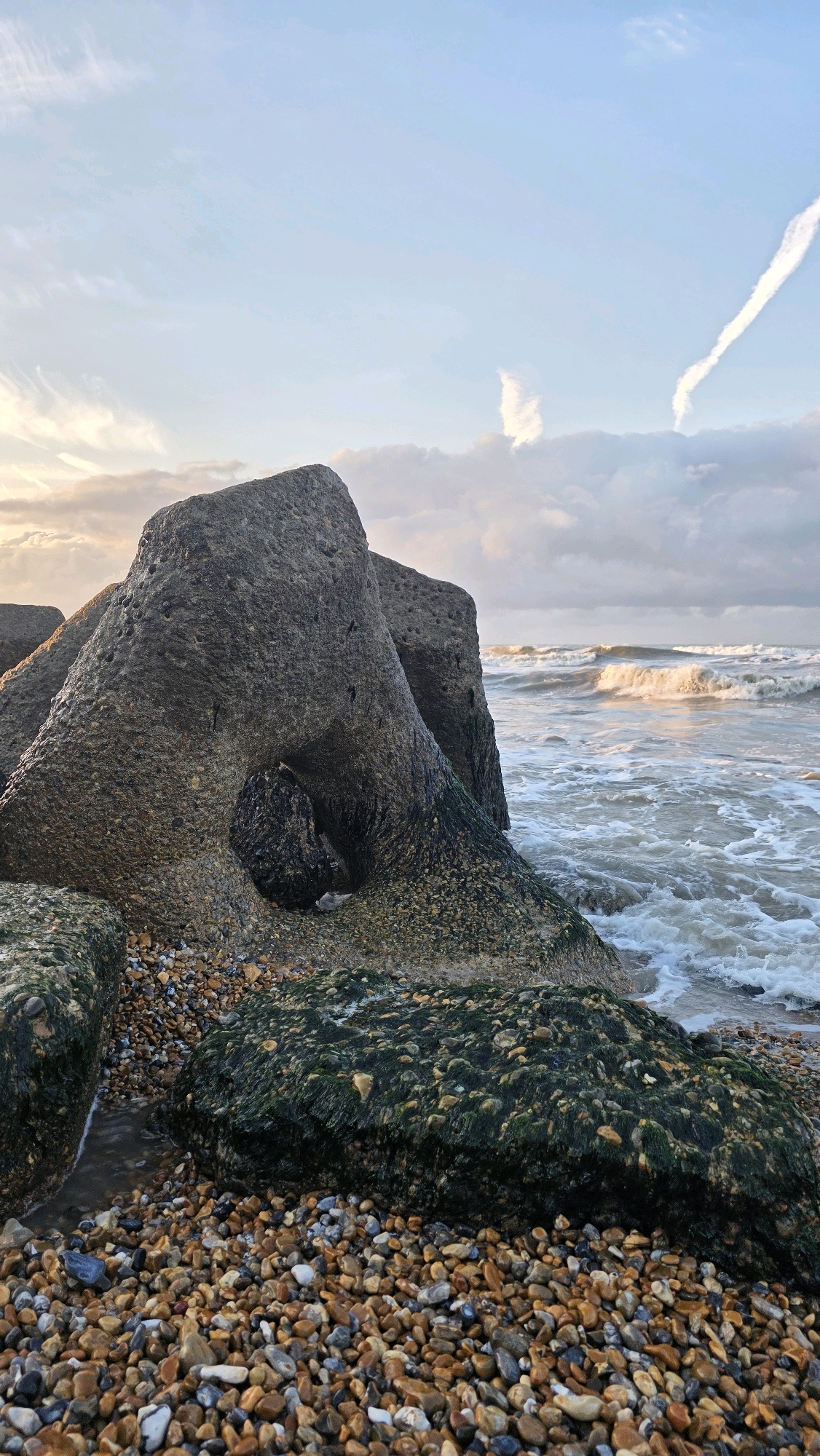 A naturalistic, eye-level, medium-long shot captures a rocky beach at St Leonards, United Kingdom, under a pale blue sky with wispy white clouds and two faint contrails. The foreground is dominated by a dense carpet of smooth, multi-colored pebbles, interspersed with dark green, algae-covered rocks. In the mid-ground, large, irregular concrete or stone structures, heavily textured and weathered, rise from the pebbles. One of these structures has a large, arch-like opening. To the right, the sea crashes onto the shore, creating white foam and choppy waves that reflect the soft light of the sky. The overall scene is one of a rugged coastline, bathed in the gentle light of either dawn or dusk.