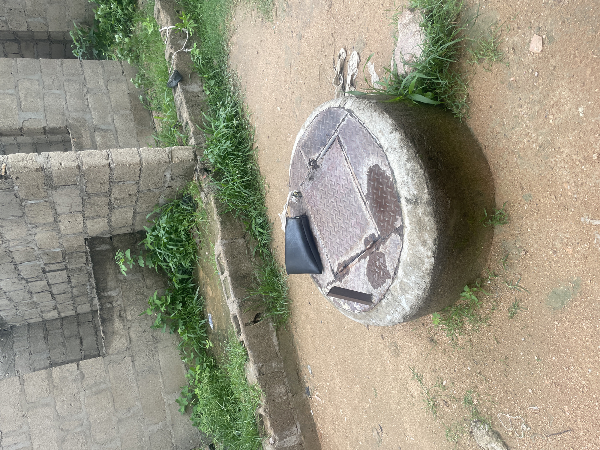 The image depicts an outdoor scene in Jalingo, Nigeria. The foreground is dominated by a circular, metal manhole cover set within a concrete rim. The manhole cover is somewhat rusty and appears wet, possibly from recent rain, with water pooled around its edges. A black handbag rests on the edge of the manhole cover.
To the left of the manhole, there is a rudimentary structure made of concrete blocks, possibly stairs or the foundation of a building, with a few small plants growing between the blocks. Beyond this structure, lush green grass grows abundantly along a raised concrete border.
The ground is a mixture of sandy earth and small pebbles, with patches of sparse vegetation. In the background, more concrete block construction is visible, also interspersed with greenery. A pair of flip-flops are placed on the ground near the manhole.
The scene appears to be set during the daytime, with natural light illuminating the area. There are no people actively engaged in any activity, giving the impression of a quiet or abandoned setting. The overall atmosphere is somewhat unkempt, with evidence of both construction and natural overgrowth. No visible text is present in the image.