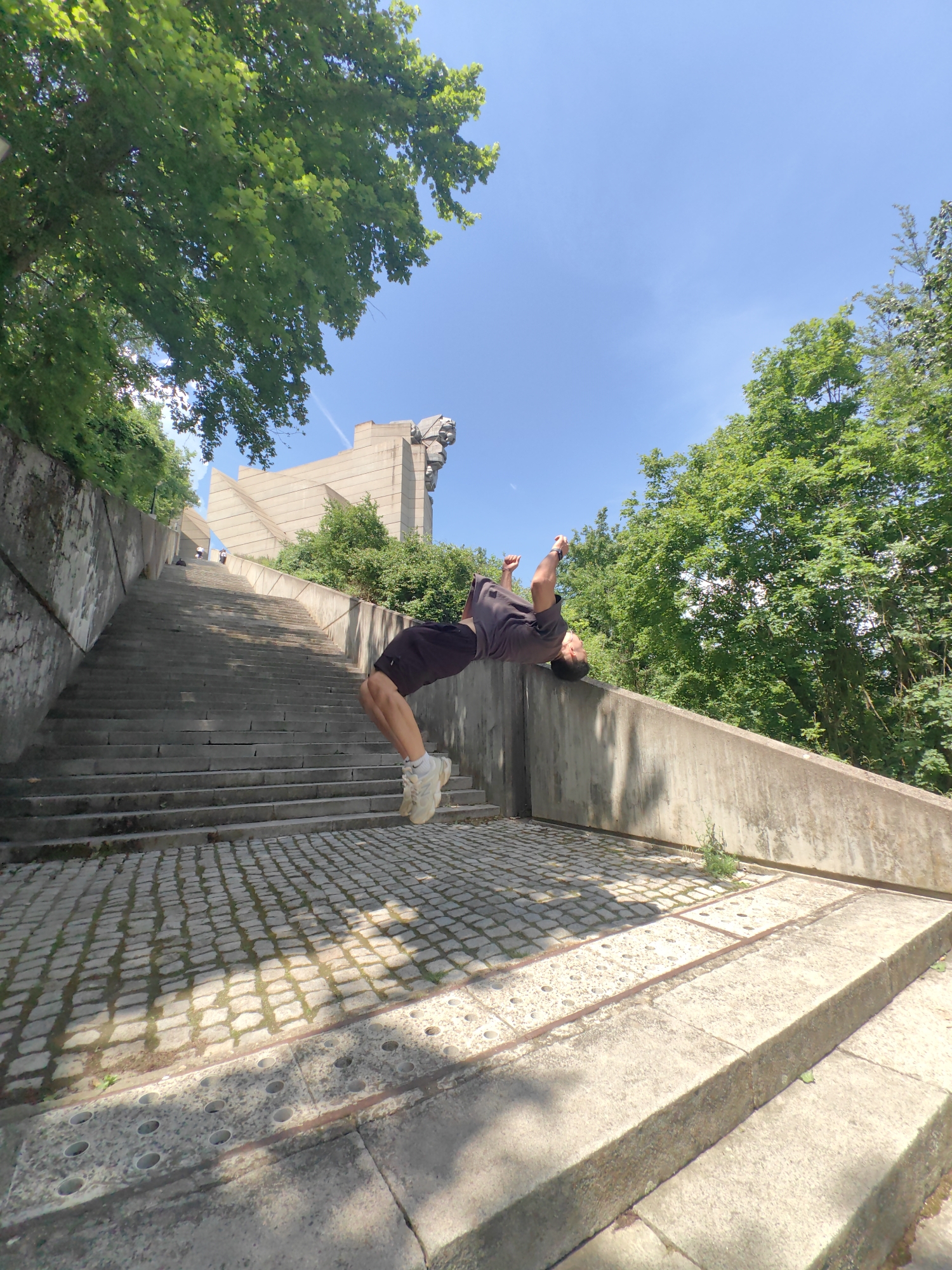 A man is performing a backflip on the stairs leading to the Founders of the Bulgarian State Monument in Shumen, Bulgaria. He is wearing a dark t-shirt, dark shorts, and light-colored shoes. The monument is visible in the background, along with a long set of concrete stairs. The stairs are flanked by concrete walls and trees. The sky is blue with scattered clouds.