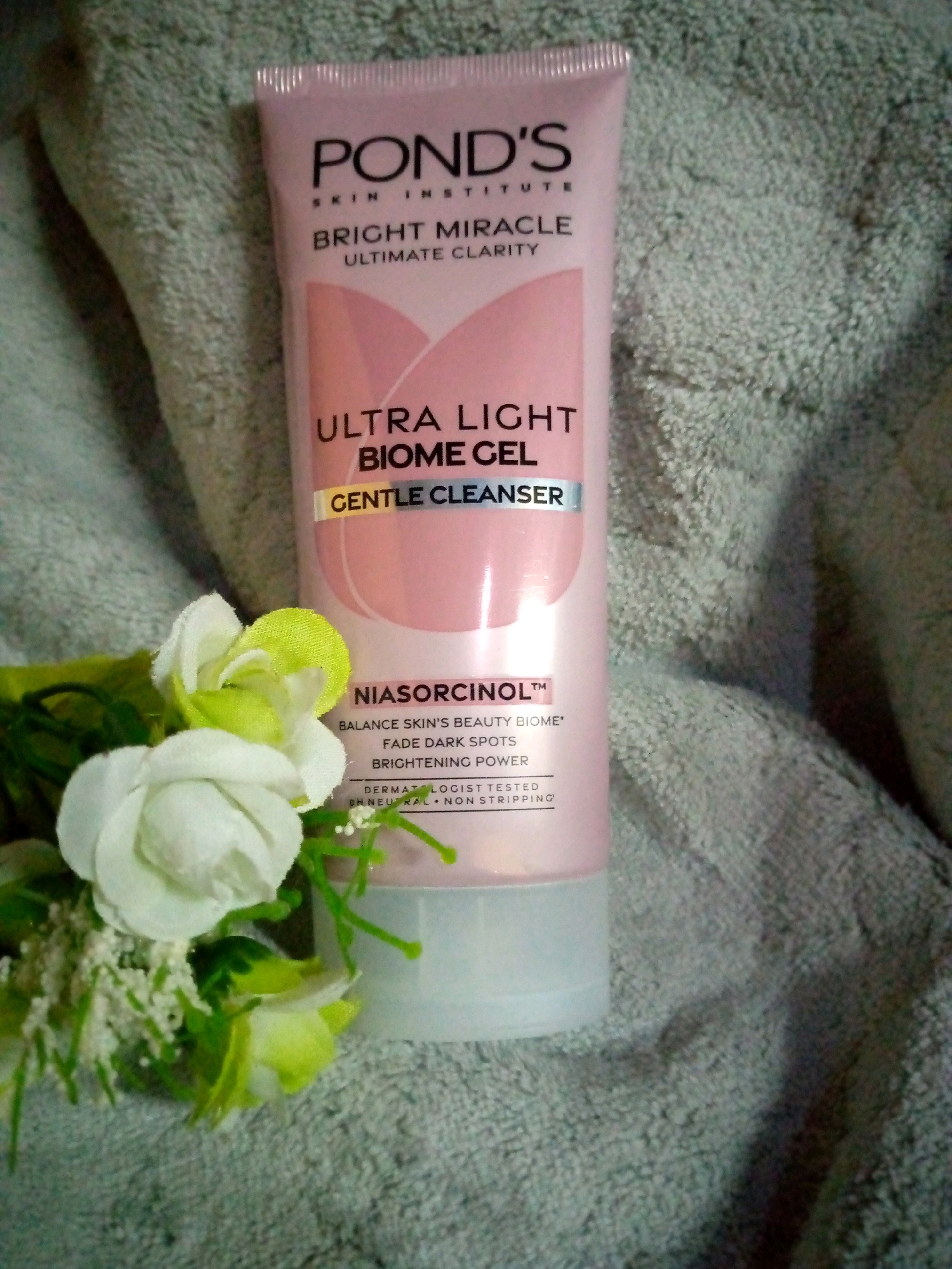 An overhead, medium shot captures a pink Pond's Bright Miracle Ultra Light Biome Gel Gentle Cleanser positioned vertically on a textured, grey towel. To the left of the bottle, a small, artificial arrangement of white and light green roses with delicate white accents is partially visible. The bottle's label prominently displays the Pond's logo, "SKIN INSTITUTE," "BRIGHT MIRACLE," "ULTIMATE CLARITY," "ULTRA LIGHT BIOME GEL," and "GENTLE CLEANSER" in various fonts and colors. Below "BIOME GEL," a strip reads "NIASORCINOL™," followed by text detailing its benefits: "BALANCE SKIN'S BEAUTY BIOME," "FADE DARK SPOTS," and "BRIGHTENING POWER." The bottom of the label includes smaller text: "DERMATOLOGIST TESTED," "PH NEUTRAL," and "NON STRIPPING." The towel's textured surface creates a soft, diffused background. The lighting is low, casting subtle shadows. The scene is static and does not depict any specific location within Sumberwono, Indonesia, other than the object itself.