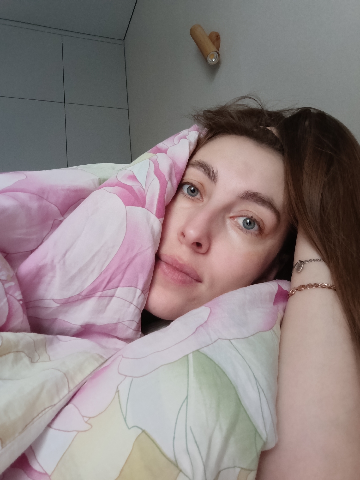 A woman is lying in bed, covered by a pink and white floral patterned blanket. She is looking directly at the camera with gray eyes, light skin, and brown hair. She wears two bracelets on her wrist, one silver and one rose gold. The setting appears to be a bedroom, featuring a wall with a modern, wooden light fixture and white cabinets. The location is likely Chaikovsky, Russia.