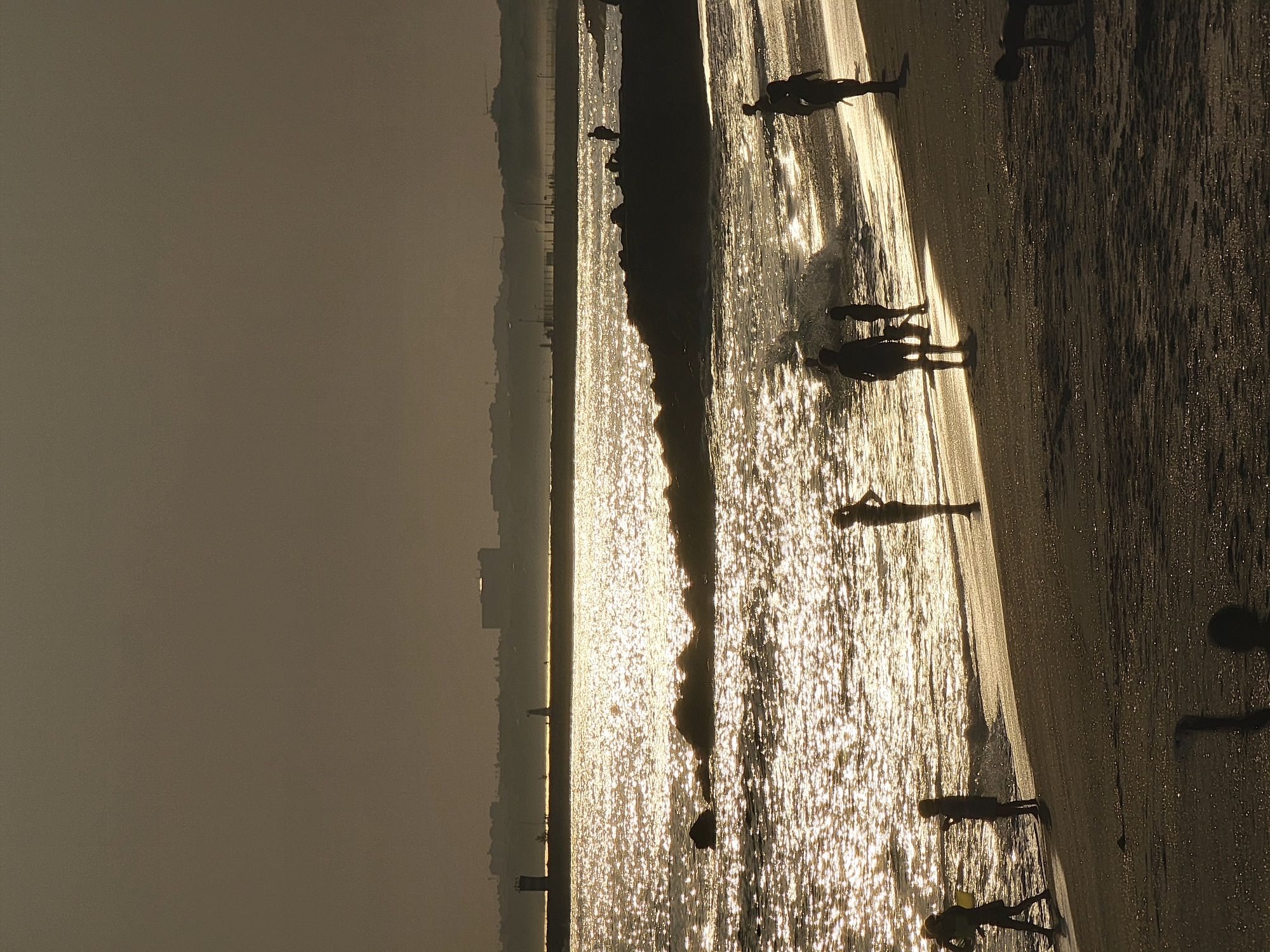 The image depicts a beach scene at sunset in Quarteira, Portugal, with silhouetted figures on the shore and in the water. The sun's rays create a sparkling effect on the ocean's surface. In the distance, a breakwater or pier extends into the sea, with a lighthouse visible on its far end. The skyline of Quarteira can be seen in the hazy background, with buildings silhouetted against the fading light. Several individuals, including children and adults, are scattered across the sandy beach and wading in the shallow water, their forms rendered as dark shapes against the bright, shimmering sea and sand. Some are standing, others appear to be walking or playing. The overall atmosphere is one of a peaceful evening by the coast.