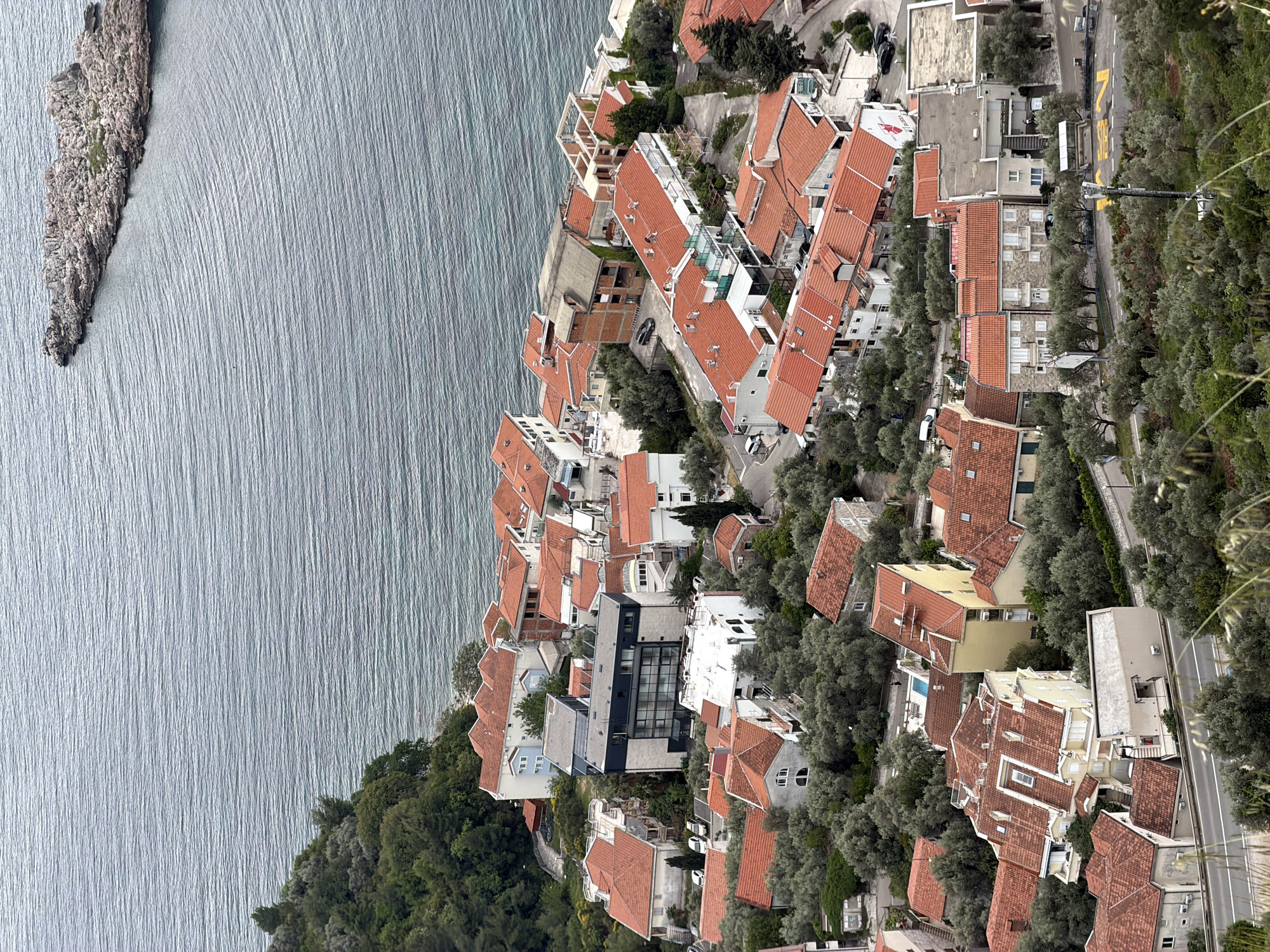 The image is an aerial view of Sveti Stefan, Montenegro, showing red-roofed buildings nestled amongst greenery along the coastline next to a body of water, possibly the Adriatic Sea. A road runs parallel to the buildings and coastline, separated by trees. An island is visible to the left.