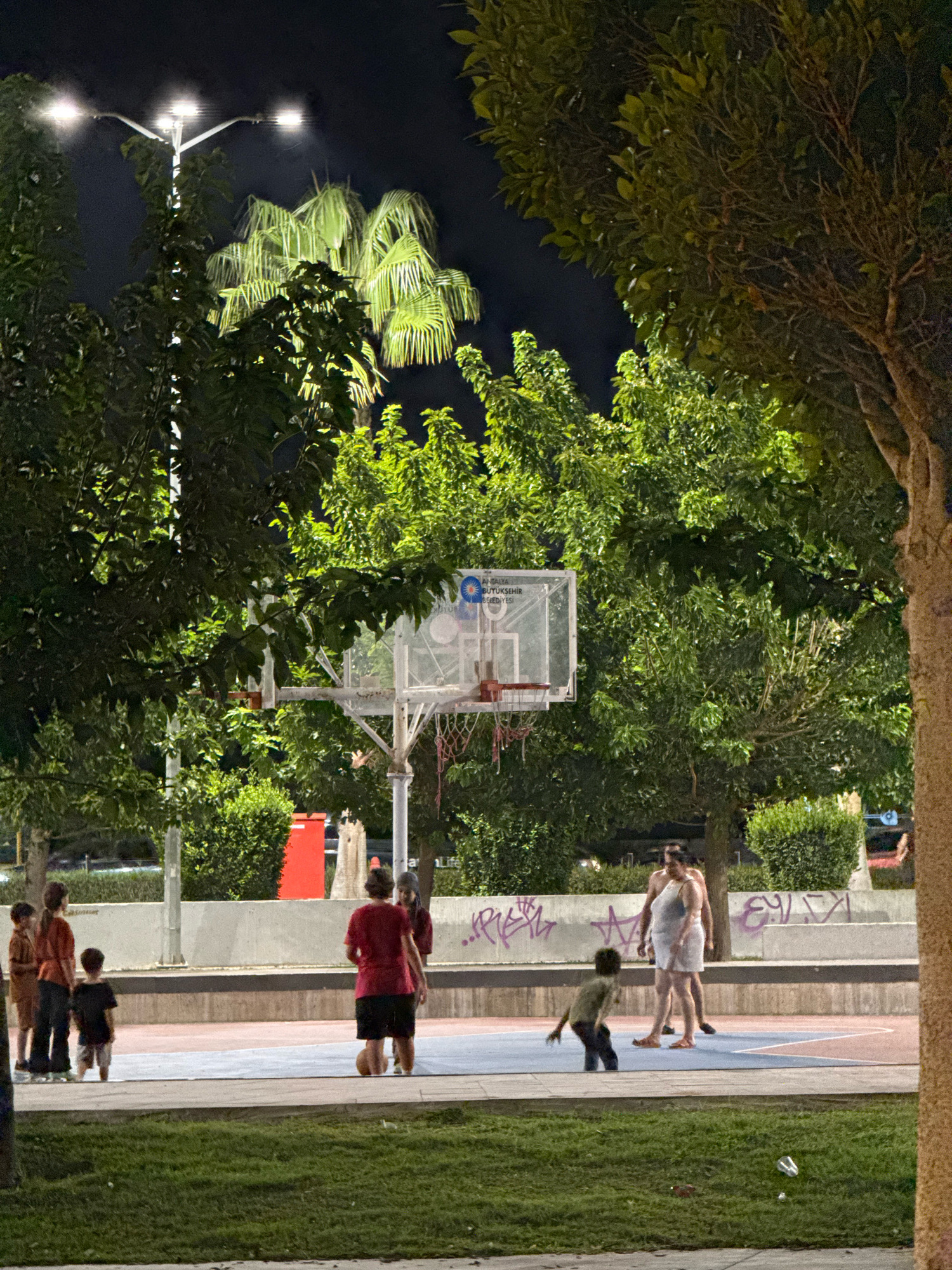 The image depicts an outdoor basketball court at night in Antalya, Turkey, illuminated by streetlights. Several individuals, including children and adults, are present on or near the court. In the foreground, a person is playing basketball. Further back, a group of children are on the court, some appearing to be watching or playing. Two adults are standing near the basketball hoop. The court is surrounded by trees and vegetation, with a wall in the background displaying graffiti. A plastic bottle and some litter are visible on the grass in the lower right corner. The overall scene suggests a public recreational area at night.
