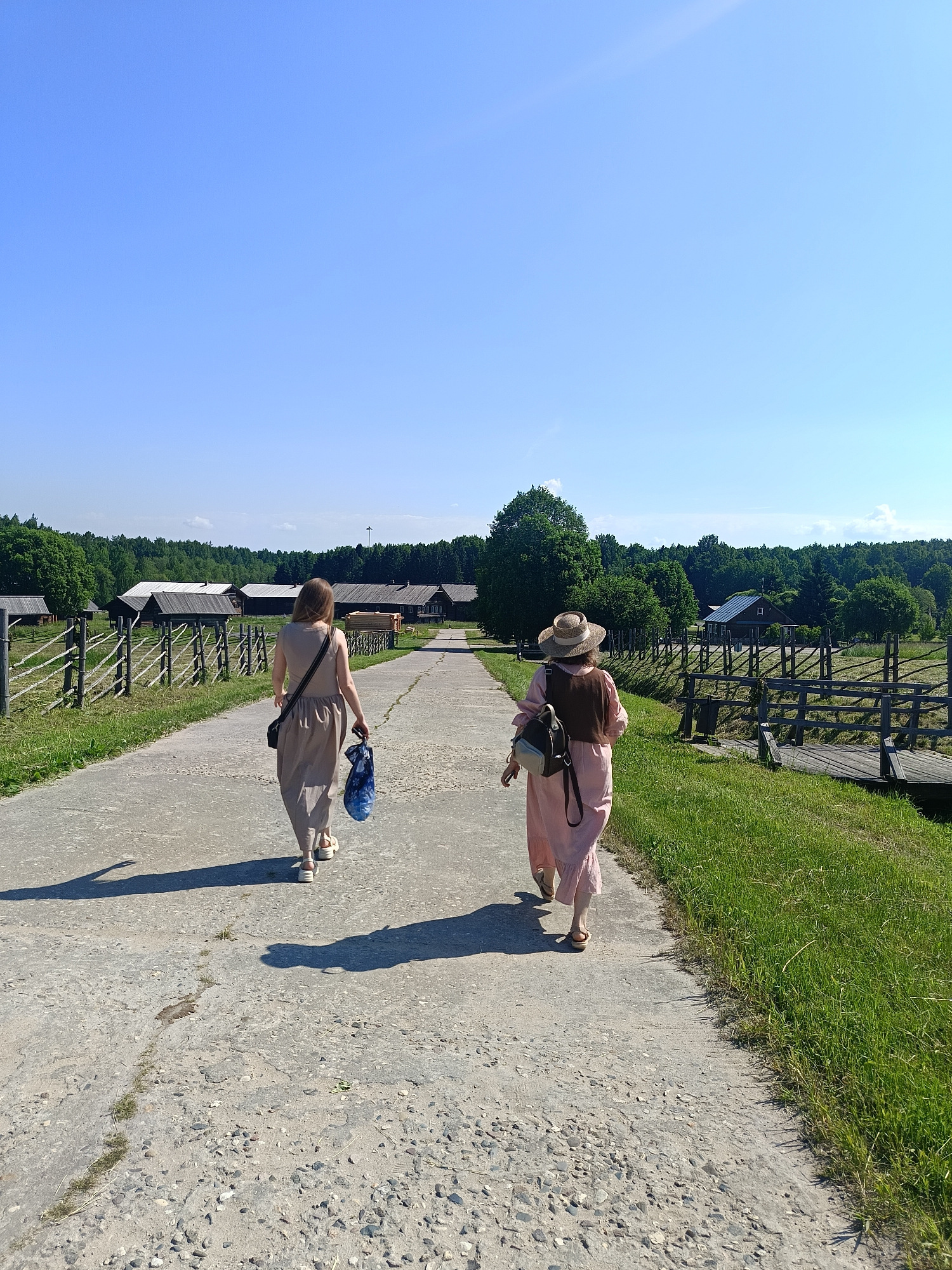 Two women are walking away from the camera on a paved path in a rural setting, likely in Popovska, Russia. The woman on the left is wearing a light-colored, floor-length dress and carrying a blue patterned bag in her right hand. The woman on the right is wearing a straw hat, a pink dress, and a brown vest, and is carrying a backpack. They are walking towards a cluster of wooden buildings with dark roofs, surrounded by green trees and a wooden fence. The sky is clear and blue, and the sun is casting long shadows. The environment appears to be a historical settlement or village.
