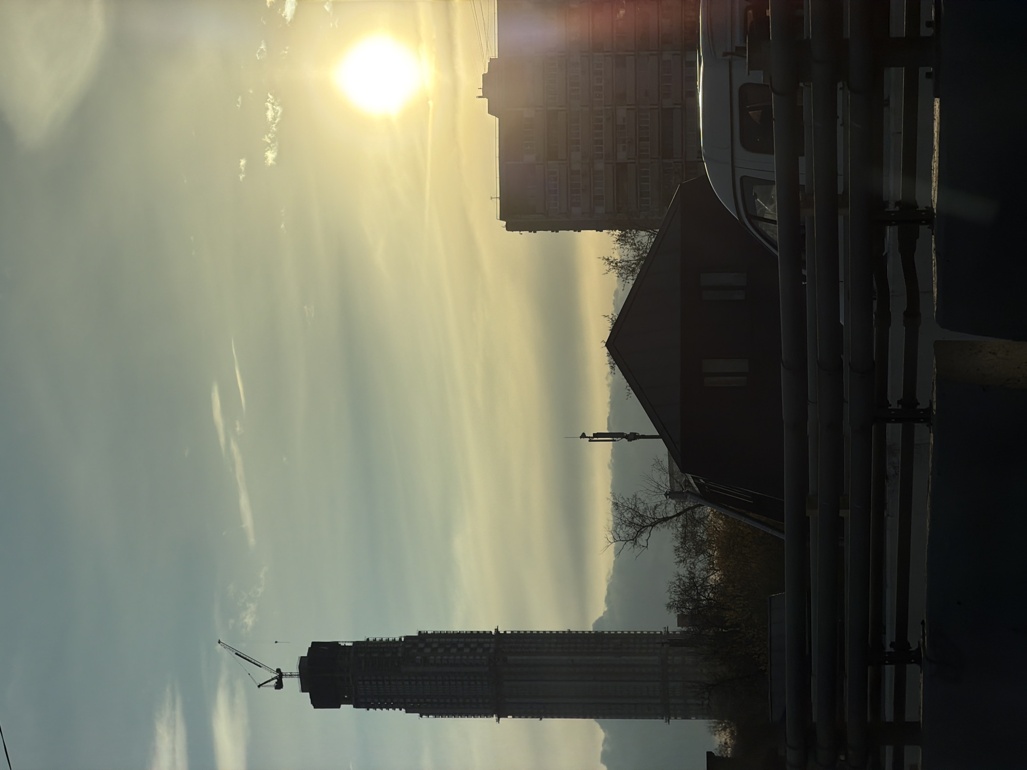 The image shows a cityscape at sunset or sunrise in Moscow, Russia. In the foreground are metal pipes and a blurred van. Behind them is a building with a peaked roof, and a larger apartment building is visible in the background. Further in the distance, a tall skyscraper under construction is visible, with a crane on top. The sky is bright with the sun shining through clouds, creating a golden haze.