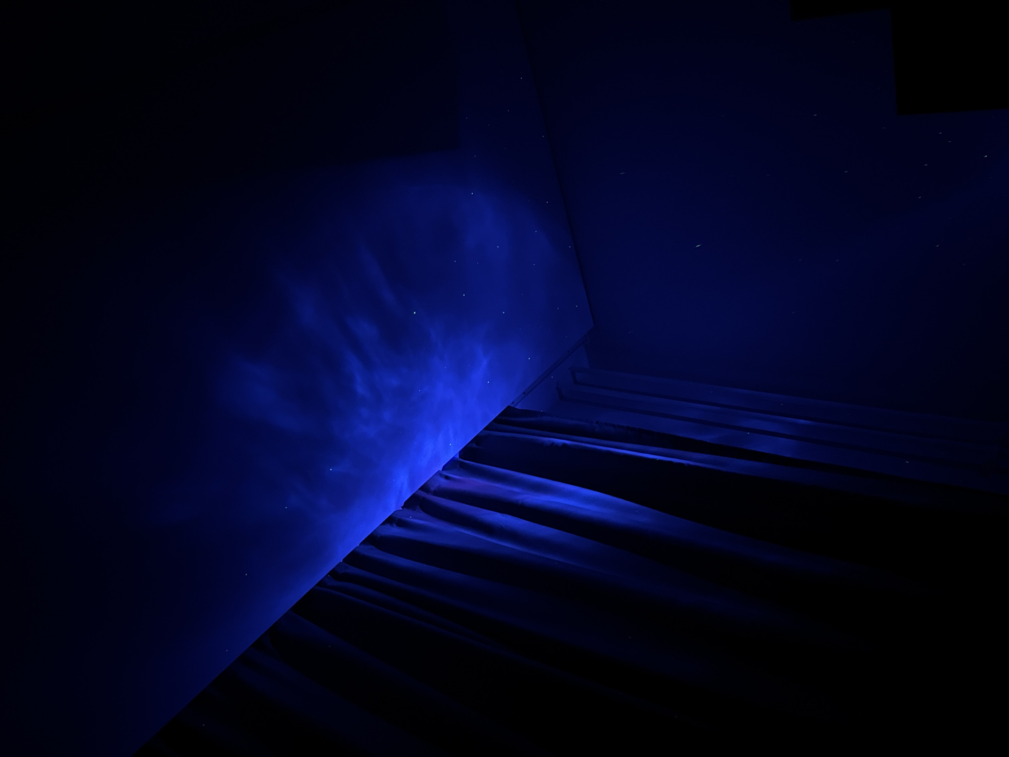 The image depicts the interior corner of a room, illuminated by a blue light source. The walls are a dark shade of blue, with a lighter, swirling blue pattern on the left wall. This pattern could be due to light projection or a decorative element. A set of stairs or a tiered structure is visible in the lower right corner, partially obscured by darkness. The lighting is dramatic, with strong contrast between the illuminated and shadowed areas. Minute details, such as dust particles or imperfections on the wall, are visible in the light. The location is not evident from the visual information.