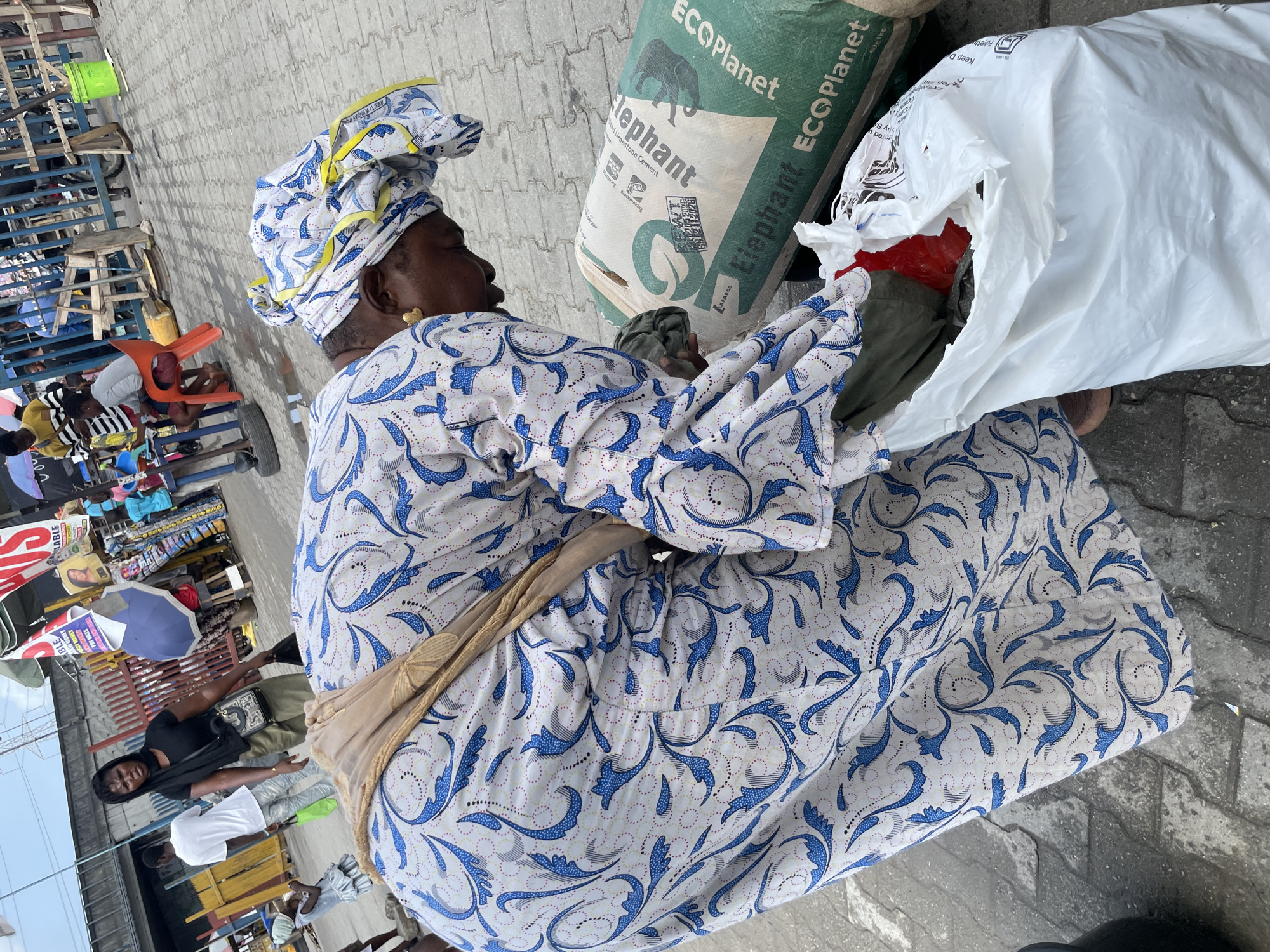 An older, dark-skinned woman wearing a colorful headwrap and a white and blue floral patterned dress sits on the gray-paved street of Lekki, Nigeria. Her dress has a beige sash around the waist. She is surrounded by objects: a large green "Elephant ECOPlanet" bag, and a large white bag. The woman's hands are inside the white bag, and there are glimpses of green and red fabric inside. In the blurry background, there is a crowd of people, some sitting on blue chairs, along with colorful signs and umbrellas.