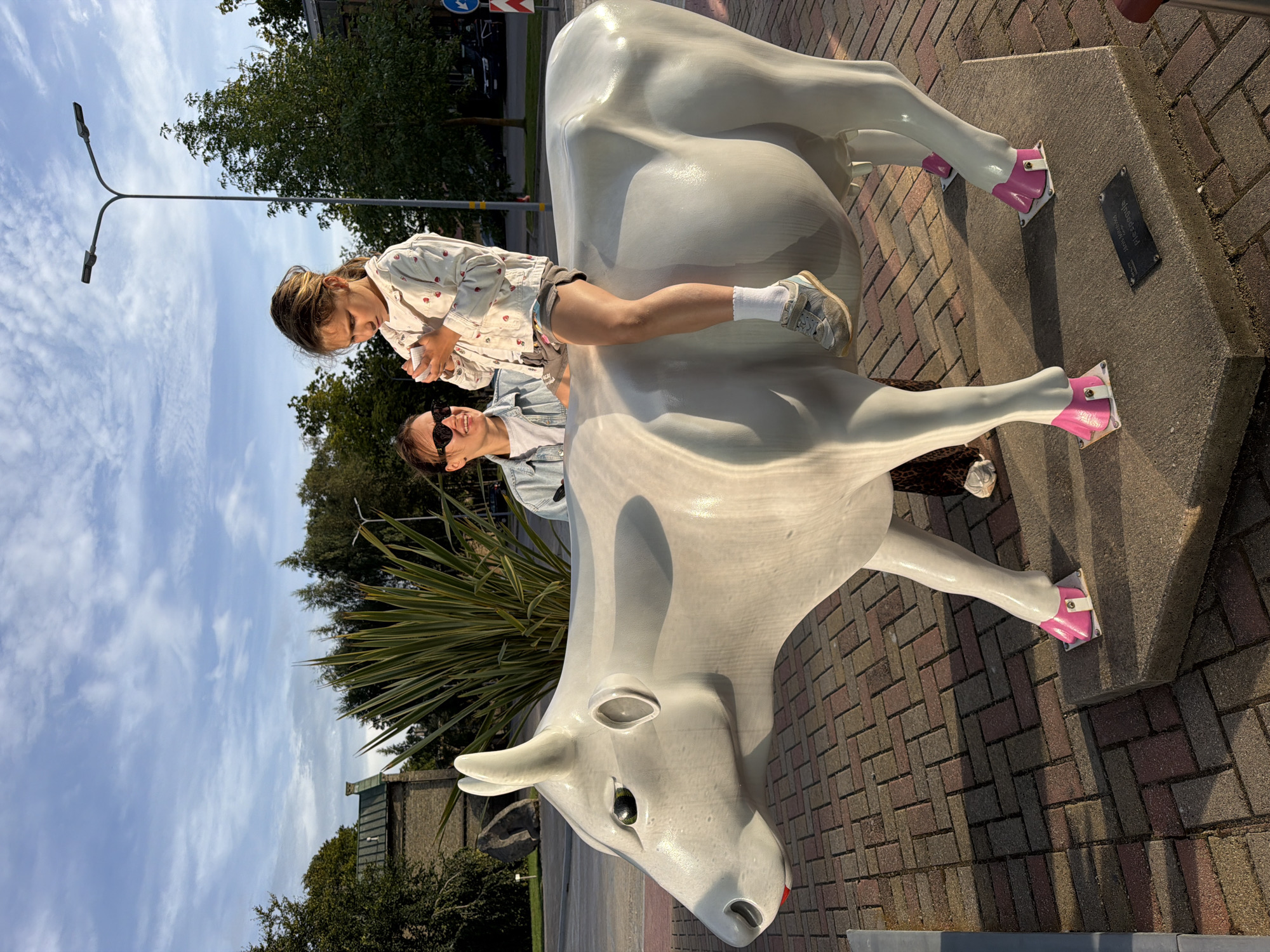 In Ventspils, Latvia, a young girl sits atop a large, white cow statue situated on a paved area with a brick-like pattern. The statue has pink accents on its hooves. The girl is holding a small, white object in her hands. Behind the statue, a woman wearing sunglasses and a denim jacket is visible. She is looking towards the girl. The environment includes trees, buildings, and a streetlamp in the background. The sky is partly cloudy.
