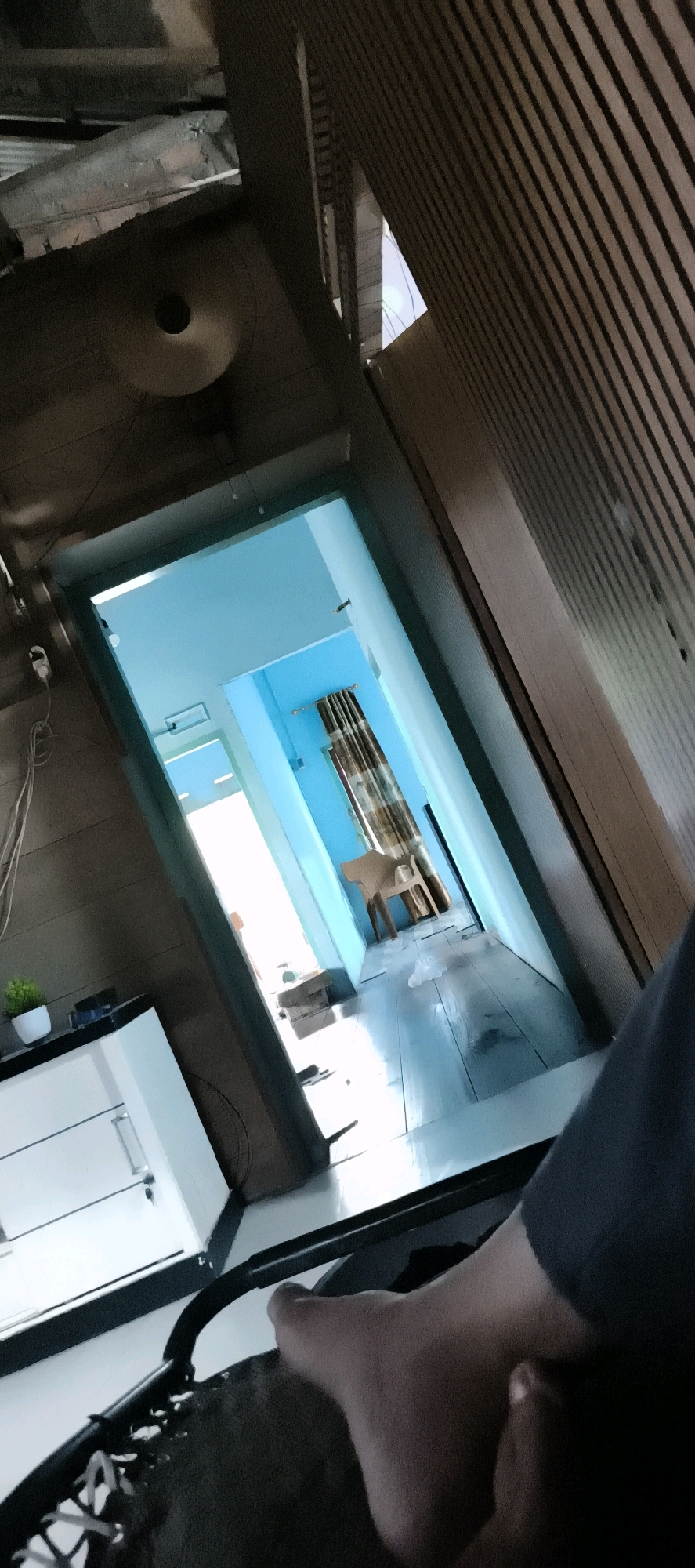 The image shows an interior view of a hallway, likely in a residential or commercial building. The hallway walls are painted a vibrant blue, and the floor appears to be made of light-colored wood planks. On the right side of the hallway, there is a wooden chair and a patterned curtain. The hallway leads to an exit or another room, where natural light is visible. In the foreground on the left, a white cabinet with drawers is partially visible, with a small potted plant on top. In the lower right portion of the image, a person's bare foot and leg are in view, suggesting they are sitting or resting. The overall impression is of a clean and possibly modern interior space. The context of Muaro Jambi, Indonesia, indicates the geographical location of this scene.