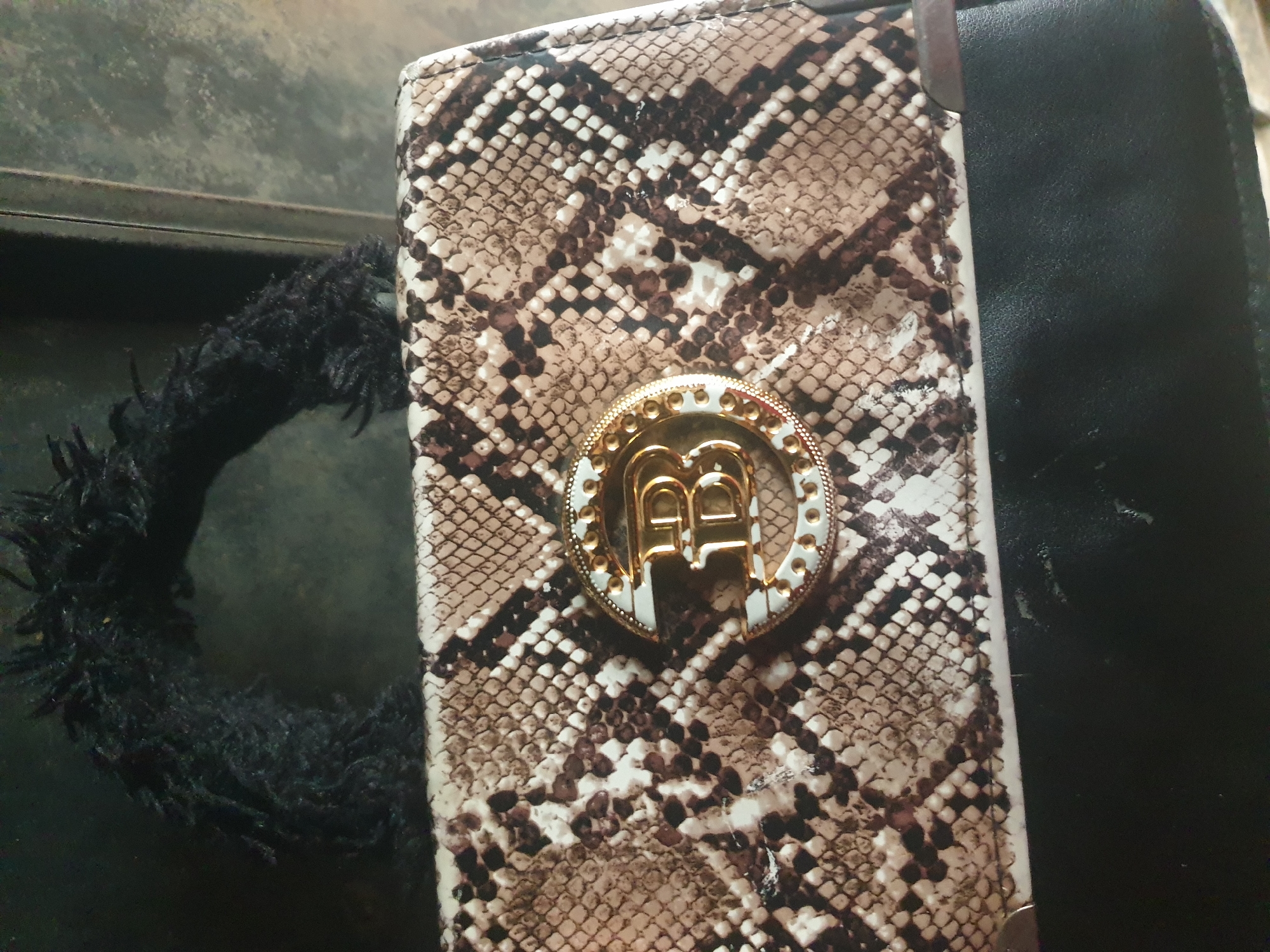 A close-up, overhead shot shows a snakeskin-patterned clutch bag with a fluffy black handle. A circular gold emblem with the letter "B" is centered on the front of the bag. The bag rests on a dark, textured surface, possibly a table or chair. The background is dark and out of focus, with a hint of a metallic vertical surface on the right side. There are no people present. The image does not provide any information to determine its location in Jalingo, Nigeria.