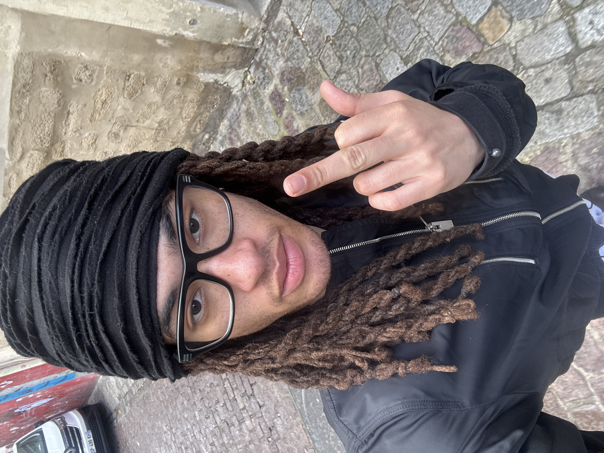 A medium-shot selfie captures a male individual on a cobblestone street in Paris, France. The subject, facing the camera, wears a black fabric headwrap, black-framed rectangular glasses, and a black zipped jacket. He has brown dreadlocks extending from beneath the headwrap. His right hand is raised in the foreground, with the middle finger extended. The street surface is paved with irregular grey and brown cobblestones. In the left background, a white Renault vehicle with a French license plate (GP-499-KH partially visible) is parked against a wall displaying red and white paint or graffiti. The right background features a rough, light-colored stone wall with visible mortar lines.