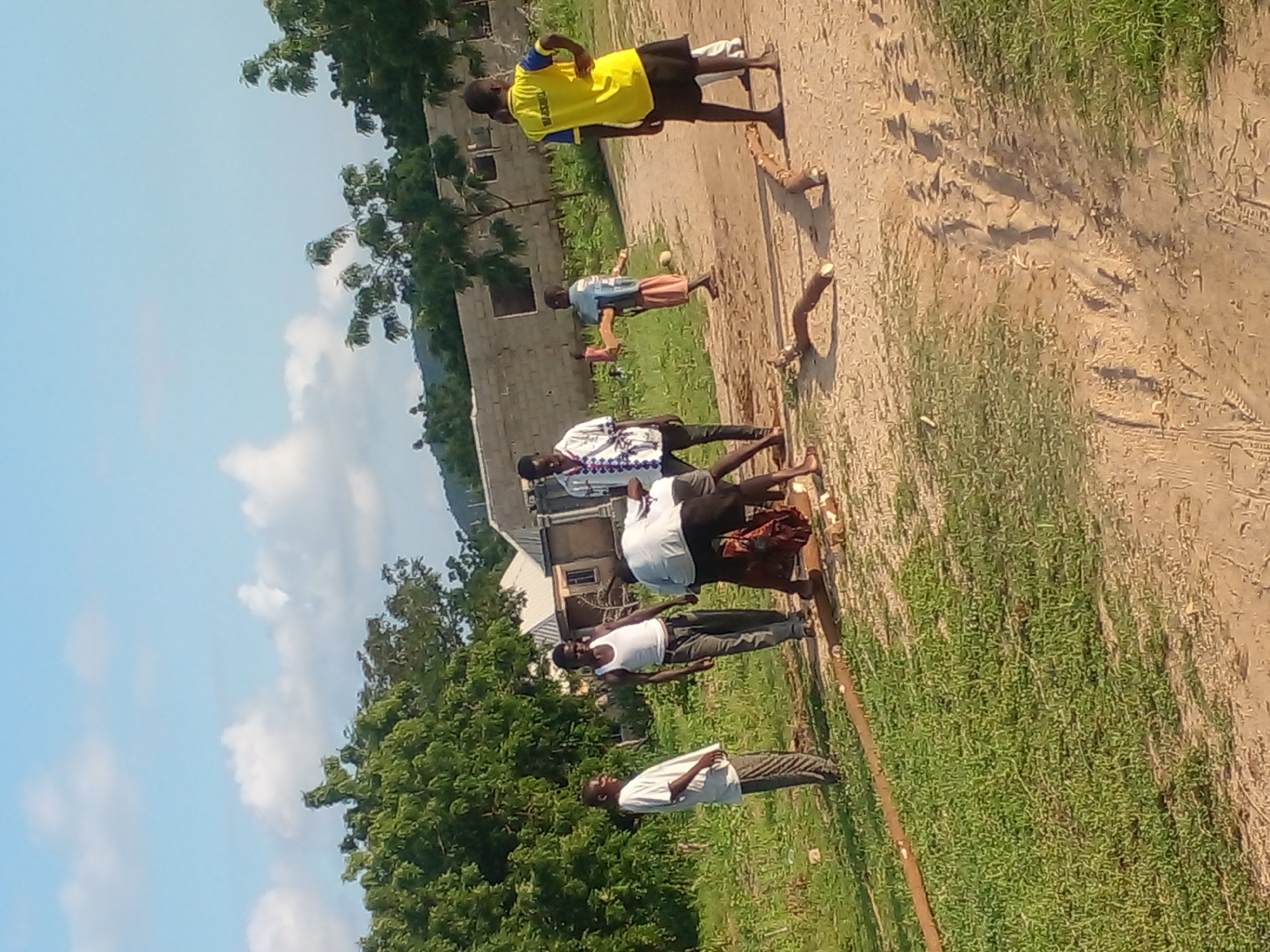 The image shows a group of individuals, possibly children and young adults, gathered on a dirt path in Langtang, Nigeria. The scene appears to be outdoors, with lush green grass lining one side of the path and trees in the background. Several structures, including buildings with brick walls and roofs, are visible in the distance, suggesting a semi-urban or rural setting.
In the foreground, a few individuals are engaged in an activity involving logs or branches lying on the ground. One person, wearing a white shirt, is bent over, seemingly working with the wood. Another individual, in a white tank top and dark pants, stands nearby, observing. A person in a yellow and blue shirt, possibly a uniform, stands with their hands on their hips, looking towards the group.
Other individuals are visible in the background, including a person in a white and patterned shirt, and a younger individual in a blue shirt and pink skirt holding what appears to be a small pink object. The overall atmosphere suggests a communal activity or a moment of shared work or play. The lighting indicates it is daytime, with the sun casting shadows on the ground.