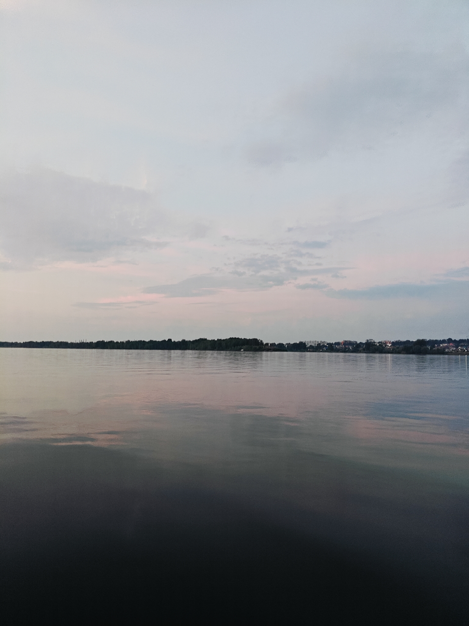 The image depicts a wide, calm body of water, likely a river or a lake, reflecting the pale, overcast sky and a distant treeline. On the far bank of the water, a dense forest stretches horizontally, with a small town or settlement visible in the distance, characterized by scattered buildings and lights. The sky is a soft gradient of pale blue and grey, with hints of pink and white clouds, suggesting either dawn or dusk. The water's surface is mostly smooth, with subtle ripples, and its color ranges from a dark, almost black hue in the foreground to lighter tones closer to the shore, reflecting the sky's muted colors. The scene is tranquil and natural, with no visible people or specific landmarks that would definitively place it in Noviy, Russia, beyond the general context provided.