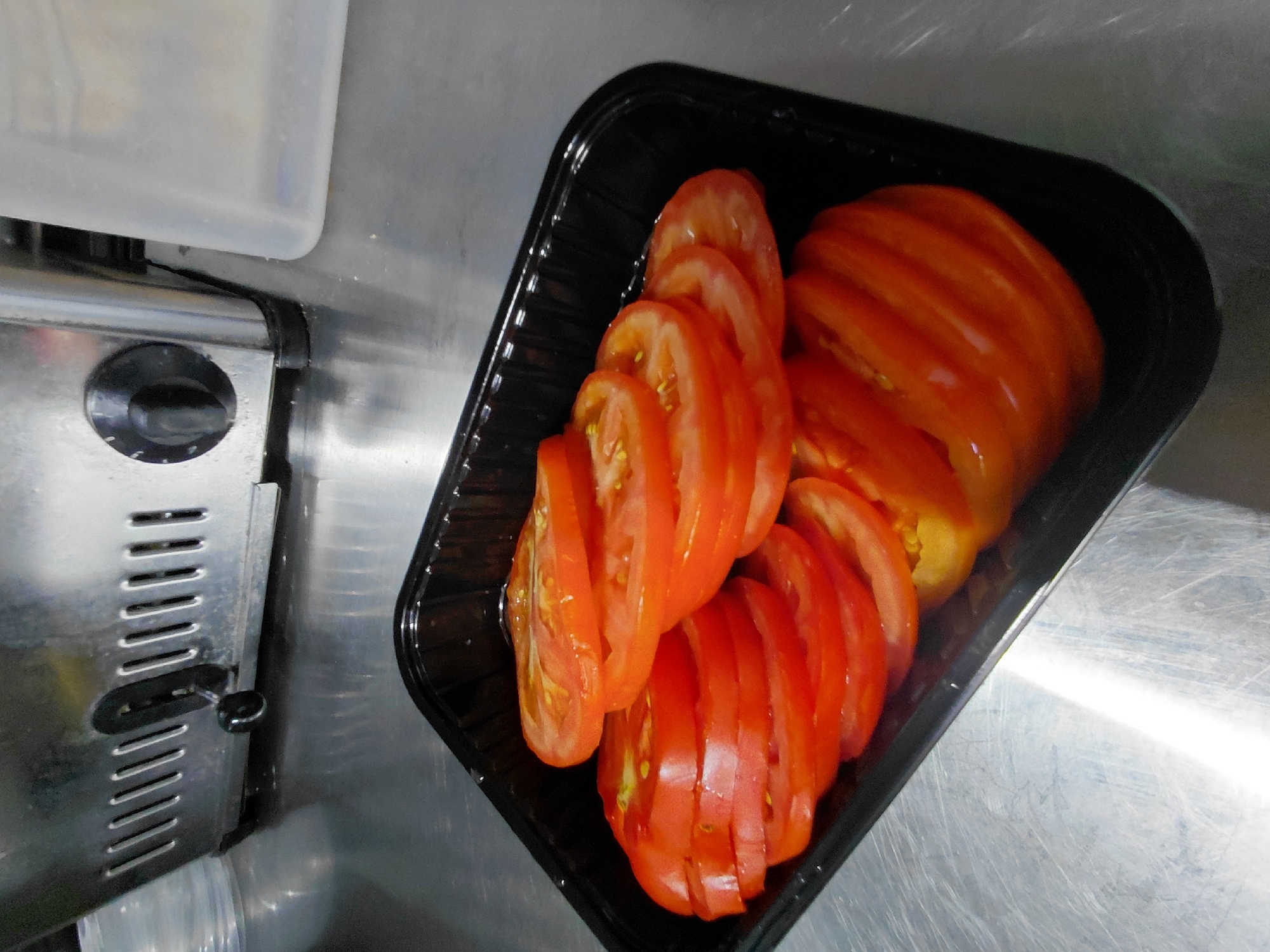A black plastic container holding sliced tomatoes is on a metallic surface. The container is filled with multiple layers of red tomato slices, with some slices partially obscuring others. The metallic surface appears to be a countertop or table, exhibiting some scratches. In the background, a stainless steel appliance with ventilation slots and a black dial is visible, along with a portion of a translucent plastic container. The scene is indoors, likely in a commercial kitchen or food preparation area, in Aktau, Kazakhstan.