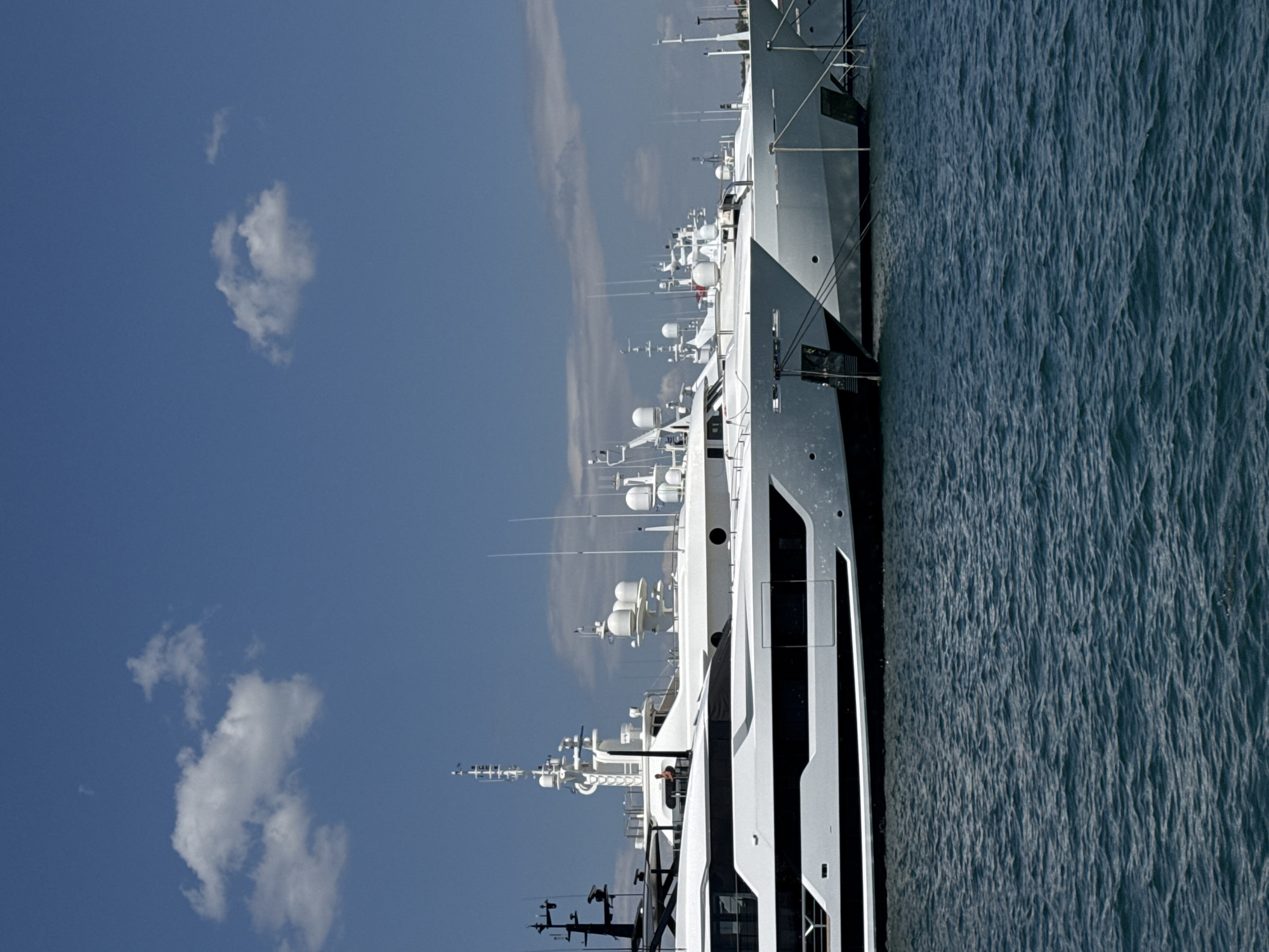 The image shows a line of large white yachts docked in the water, likely in the port of Moschato, Greece, given the context. The sky is a clear blue with a few scattered white clouds. The yachts are modern and luxurious, with multiple decks and antennas visible. The water is a deep blue and appears slightly choppy. There is at least one person visible on one of the yachts, standing on an upper deck. The overall scene suggests a wealthy and upscale environment.