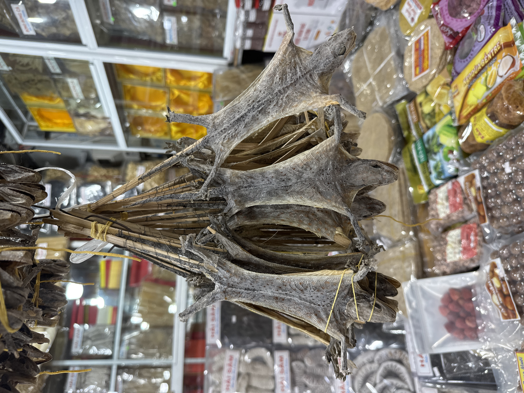 The image shows a stack of dried geckos for sale, tied together with twine, in what appears to be a market stall. The background shows other packaged food items. The location is likely in the city of Nha Trang Ward, Vietnam, as that is where the image was taken.