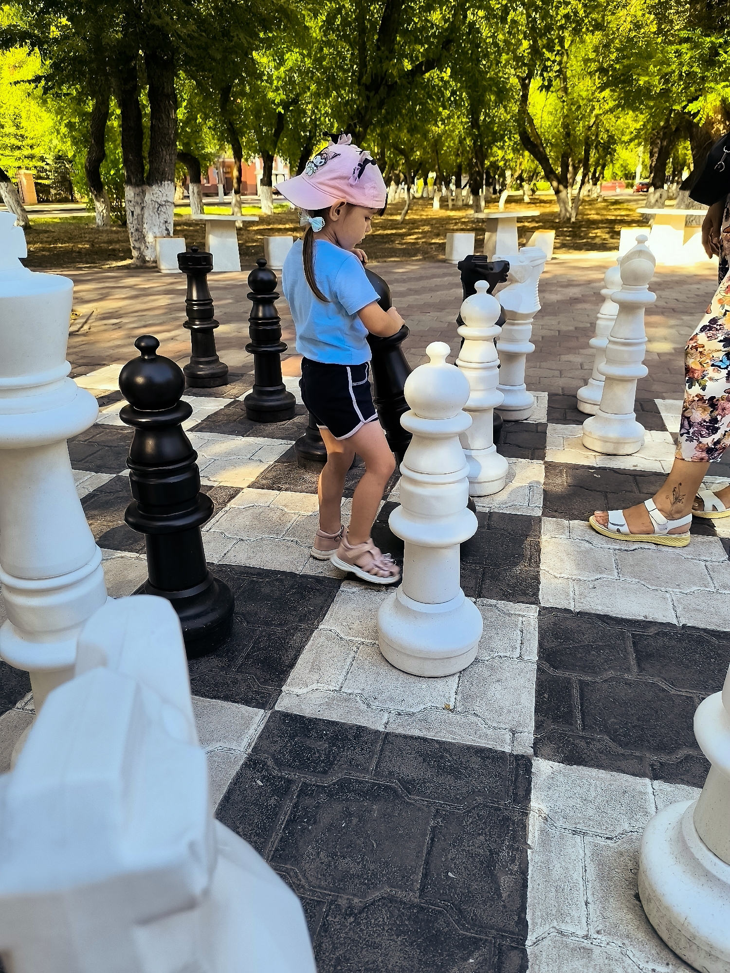 A young girl wearing a pink hat and blue shirt stands on a giant chessboard in an outdoor park setting. She is looking down, possibly at the chessboard. The chessboard is made of large black and white square tiles. Several oversized chess pieces, both black and white, are positioned on the board. In the background, there are trees with green foliage and some white benches and tables. Another person's legs, wearing a floral pattern, are visible on the right side of the image, suggesting they are also participating in the game. The overall impression is a playful scene in a park.