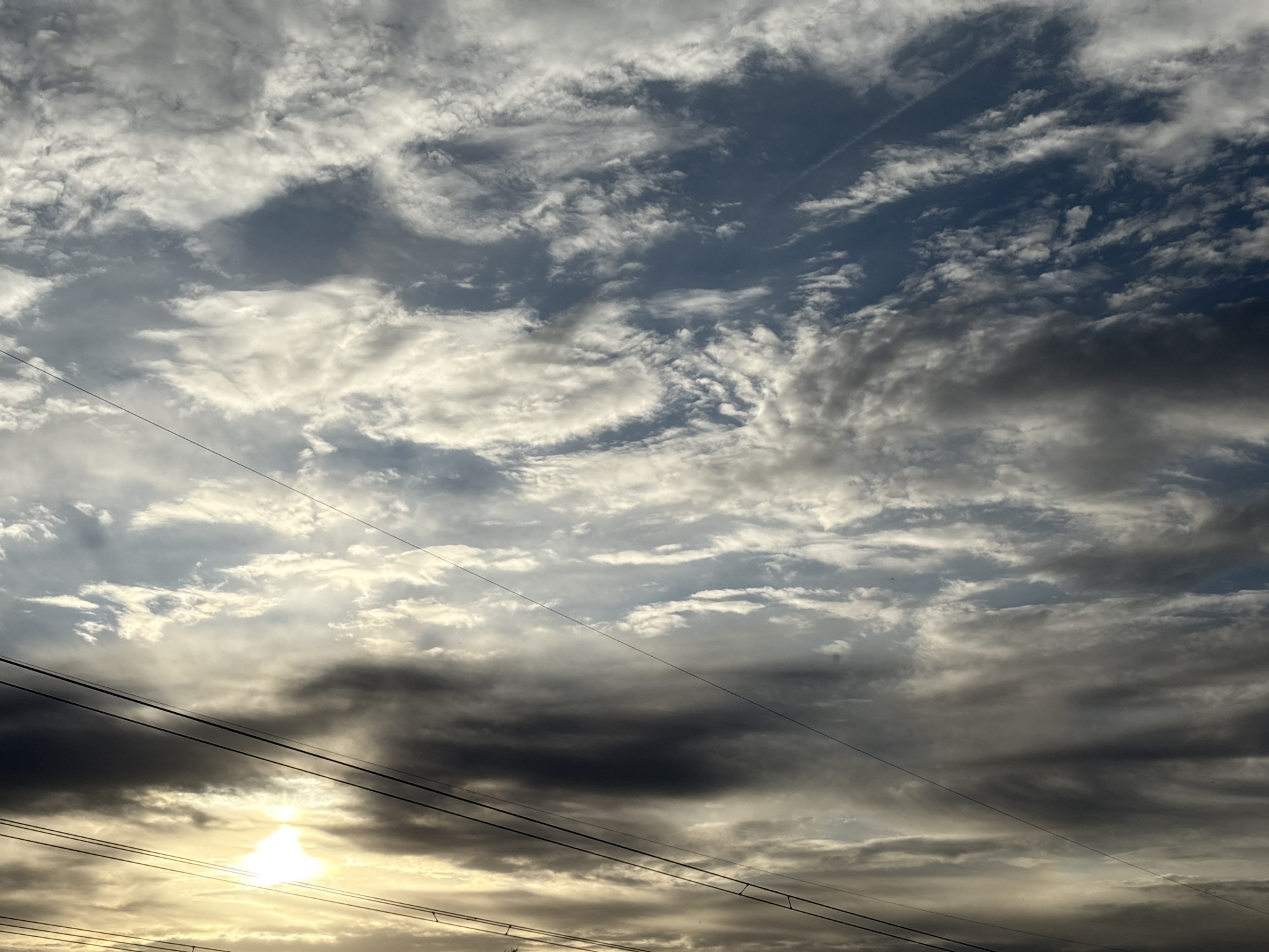 The image shows a sky filled with a mix of altocumulus and stratus clouds, with some areas of blue sky visible. The clouds range in color from bright white to dark grey, suggesting varying thickness and density. There are contrails visible in the upper right portion of the sky. In the lower part of the image, power lines are seen silhouetted against the bright light from the setting or rising sun, which is partially obscured by the clouds. The sunlight creates a radiant effect where it breaks through the cloud cover. The location is specified to be in the city of Chaikovsky, Russia, but there are no visible landmarks that would confirm this location.