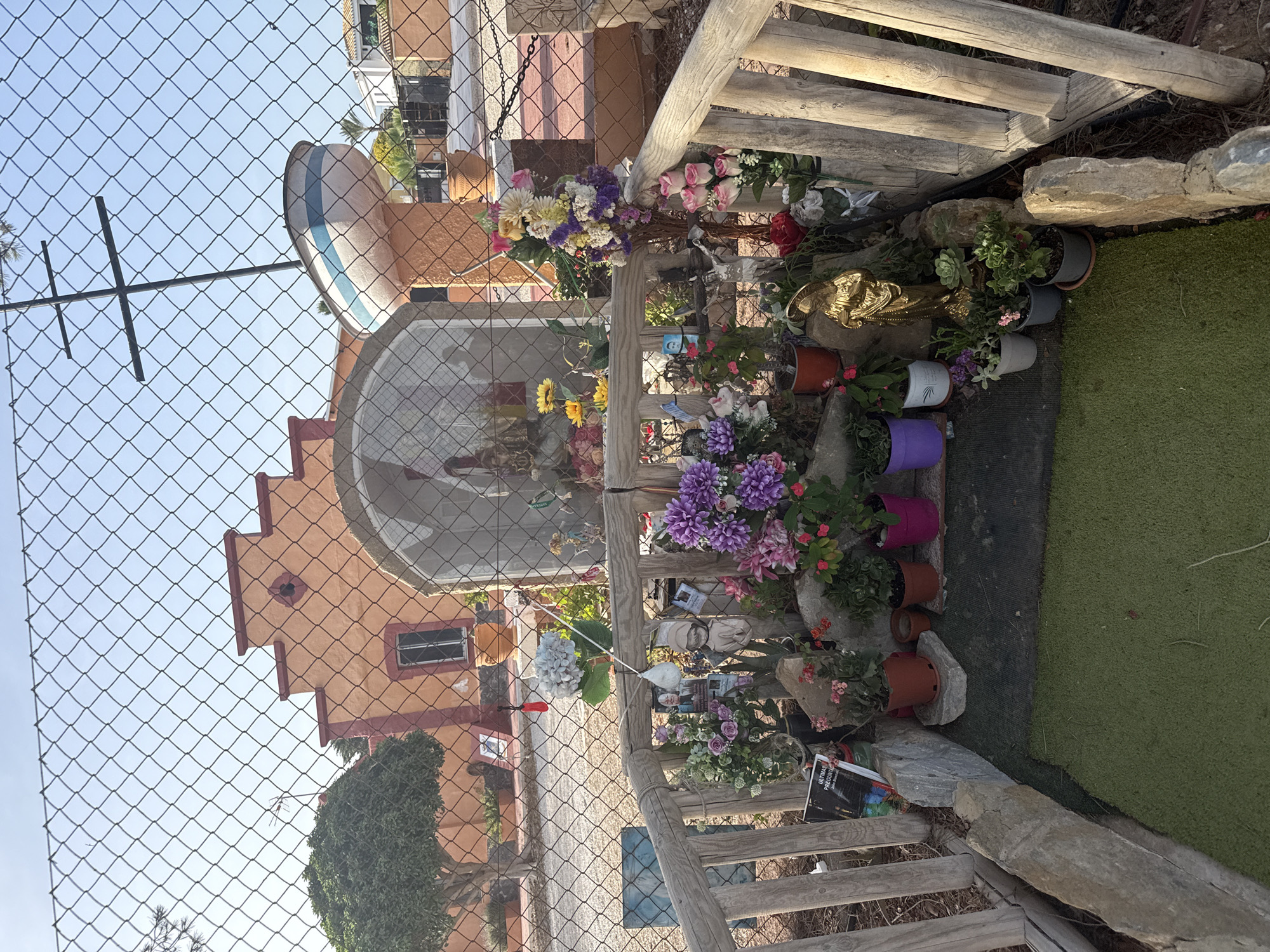 This image shows an outdoor shrine, likely in a residential area given the buildings in the background. The shrine is enclosed by a wire mesh fence. In the foreground, there is a small, green, artificial turf area bordered by rough-hewn stones and wooden fencing. A variety of potted plants with colorful flowers, as well as some succulents, are arranged on and around the shrine. There are also several decorative items, including a golden statue of a robed figure, a statue of Jesus on the cross, and what appear to be photographs or religious images. Behind the shrine, a building with orange and red trim is visible, with a small tree to its left. To the right of the building, a boat-shaped structure with blue and white stripes is mounted on a pole, topped with a cross. The sky is clear and blue, suggesting a sunny day.