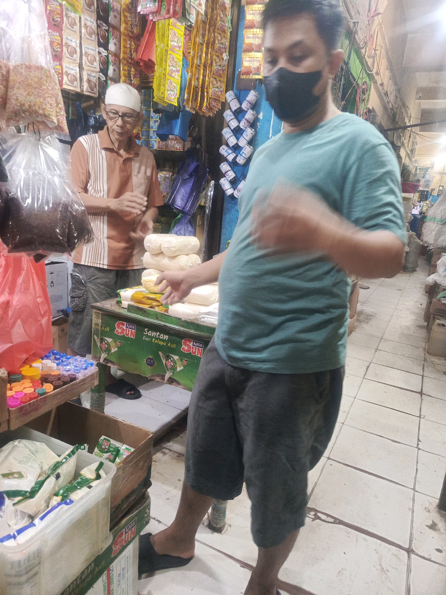 The image depicts a market stall in Kendari, Indonesia, likely within the Wua Wua Market or a similar local marketplace. Two men are present. The man on the left, wearing a white cap, glasses, and a striped polo shirt, appears to be a vendor. He is standing behind a stall filled with various goods, including bags of what appear to be food items, small colorful containers, and some larger sacks of produce. He is reaching forward, possibly to serve a customer or arrange products.
In the foreground, a second man, partially obscured and wearing a dark face mask and a teal t-shirt with dark shorts, is the focus of the image. His body is angled towards the viewer, and his right arm is blurred, indicating movement, suggesting he is interacting with the vendor or handling goods. He is standing in front of a green and white refrigerated display case labeled with "Kara SUN Santan Dari Kelapa Asli," indicating it contains coconut milk. Stacked on top of this display are several white, cylindrical packages, possibly food items.
The environment appears to be an indoor market with tiled flooring. Shelves lined with packaged goods and hanging products are visible in the background, creating a busy market atmosphere. The lighting is typical of an indoor market, with a mix of ambient and possibly artificial light sources, casting some shadows and contributing to the overall scene.
