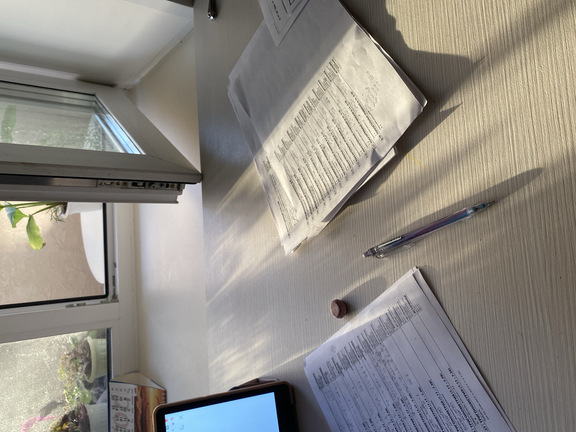 The image shows a desk with papers, a pen, a small eraser, and a tablet. The papers appear to be worksheets or study materials with mathematical problems written in Russian. Some of the topics include trigonometry and exponential equations. A window is visible to the left, with plants on the windowsill. Light is streaming through the window, casting shadows across the desk. The location is unknown; there is no indication that this is Votkinsk, Russia.