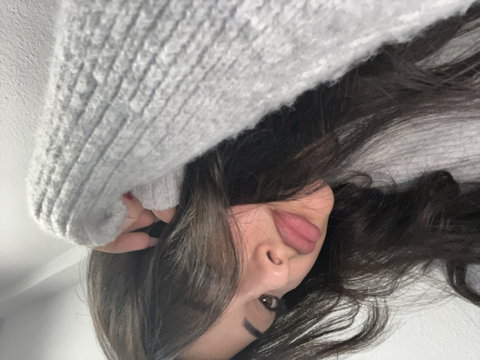 A close-up, angled portrait of an individual, likely a female, with long, dark brown hair partially obscuring the left side of her face. Her right eye, nose, and lips are visible. She wears a light grey, ribbed knit sweater. Her left hand, adorned with red nail polish, is positioned near the top of her head, with fingers visible against her hair. The background is a plain, light-colored wall.