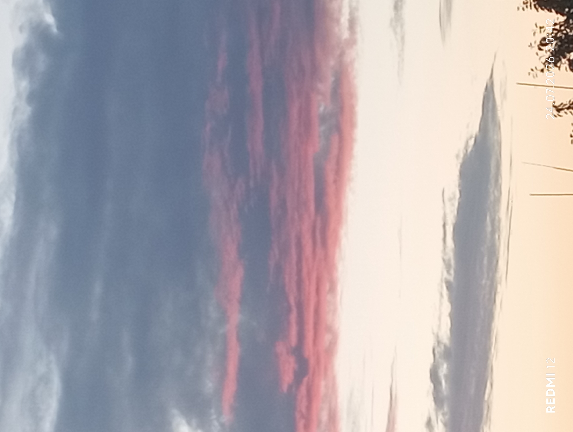 The image captures a sunset sky with layers of clouds displaying hues of gray, pink, and pale orange. The lower portion of the image shows a faint silhouette of trees against the light sky. In the bottom left corner, the text "REDMI 12" is visible, indicating the phone model used to capture the photo. In the bottom right corner, the date and time "24.07.2026 20:42" are displayed, suggesting the image was taken on July 24, 2026, at 8:42 PM. The provided location context indicates this scene is in the city of Smila, Ukraine.