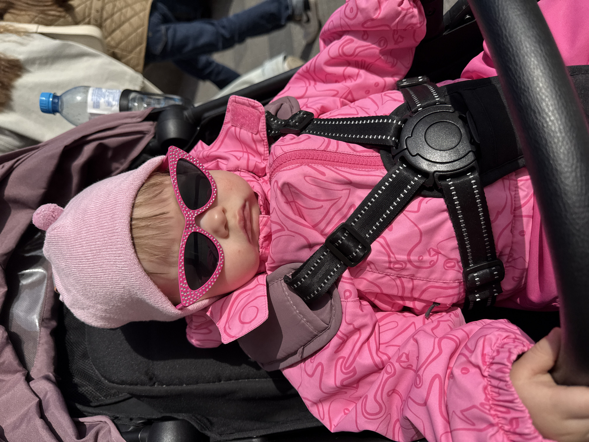 A baby wearing pink sunglasses and a matching hat and snowsuit lies in a stroller. The baby is secured with a black harness. A water bottle lies on the stroller fabric nearby. In the background, people are walking. The scene is set in an urban environment, likely Moscow, Russia.