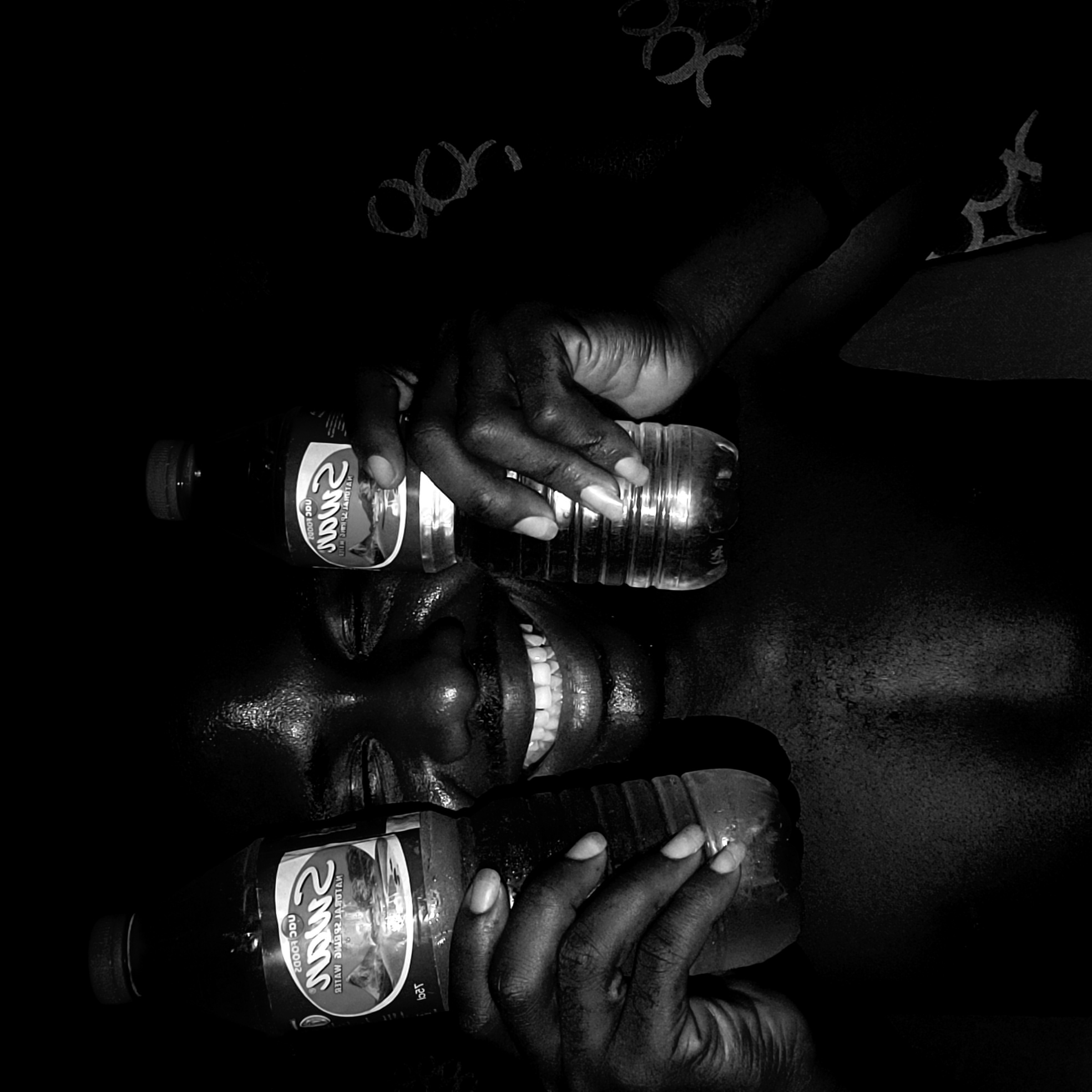The image is a black and white, rotated depiction of a person with dark skin and short nails holding two bottles of water. Their face is visible, showing a nose, partially opened mouth with teeth showing, and forehead reflecting light. The bottles appear to be partially full. One bottle label reads "SWAN" in mirrored text. The background is largely dark, with a patterned fabric or surface visible above the person's head, featuring repetitive circular designs. The image does not contain any definitive elements to confirm the location in the city of Narayi, Nigeria.