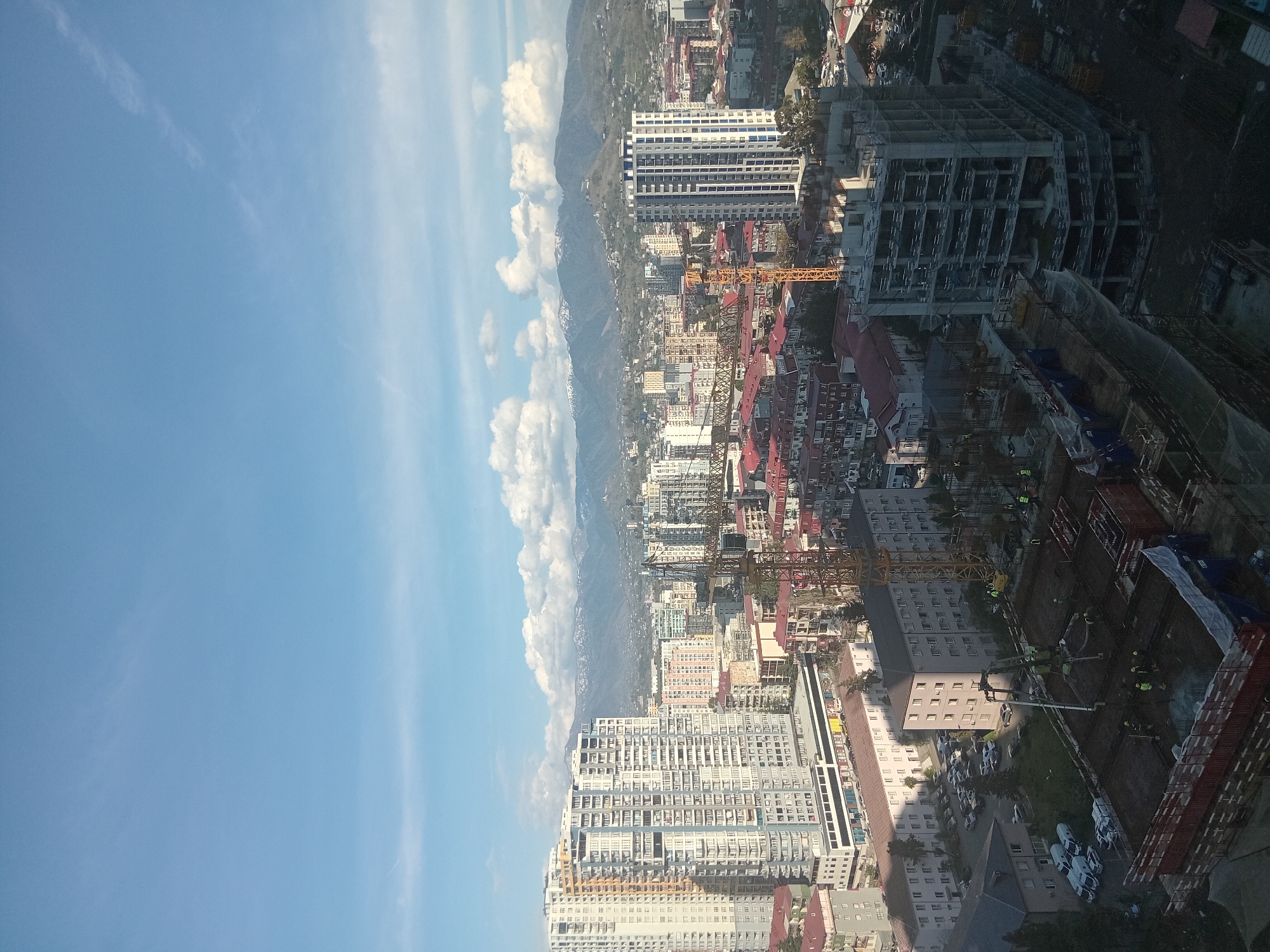 The image shows a high-angle view of Batumi, Georgia. In the background, mountains are visible, with clouds clinging to their slopes. The foreground is dominated by a dense urban landscape featuring numerous buildings, many of which are high-rise residential or commercial structures. Several construction cranes are scattered throughout the city, indicating ongoing development. The buildings show a mix of architectural styles and colors. To the right, a construction site with workers visible. The sky is blue with wispy clouds.