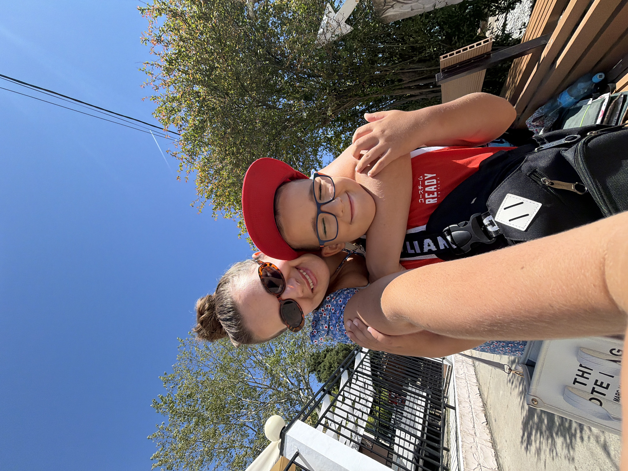 Two individuals, an adult female and a young male child, are captured in a selfie-style photograph outdoors. The female, positioned on the left, wears sunglasses and a blue floral-patterned dress, with her hair pulled up in a bun. She is embracing the child. The child, on the right, wears a red baseball cap and glasses and is dressed in a red tank top with Japanese lettering and the word "READY." He has a black backpack with a white diamond emblem. They appear to be in a relaxed and affectionate pose.
The background suggests an outdoor setting in Sochi, Russia. To the left, there is a white fence with a lamppost and the awning of an establishment. Beyond the fence, there is a pathway and foliage. To the right, lush green trees and vegetation are visible, with a portion of a wooden structure or seating area. The sky is a clear, bright blue, with power lines crossing overhead. The overall lighting indicates it is daytime, likely sunny.