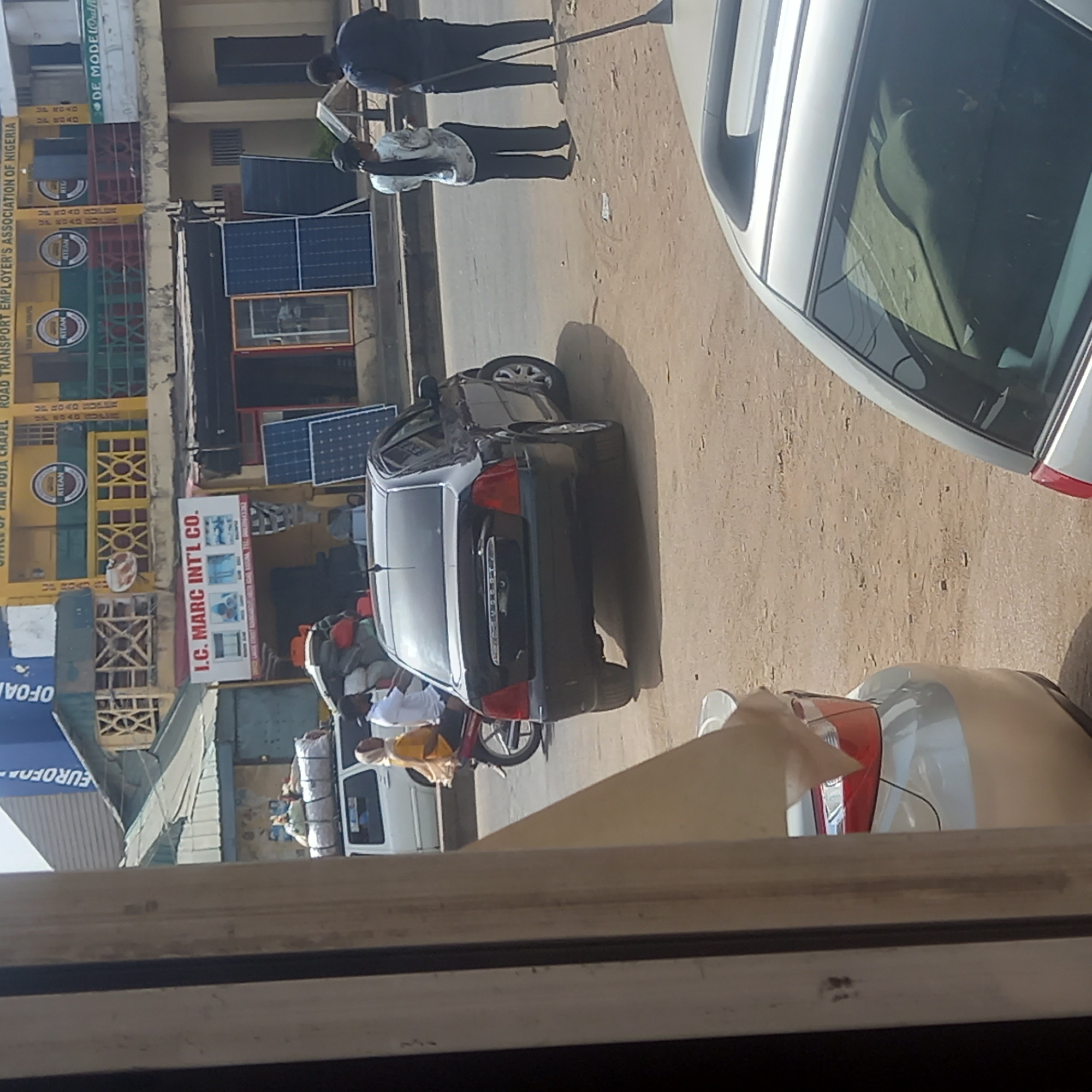 The image, taken from an elevated perspective through a window, captures a street scene in Dadi Riba, Nigeria. The street is unpaved and dusty. Several vehicles are visible: a black sedan parked on the side of the road, parts of two silver cars, and a white van partially obscured in the background. A motorcycle with two people is parked next to the black sedan. Two men are standing on the side of the road near a building. The building has several signs, one for "I.C. MARC INT'L CO." and another for "ROAD TRANSPORT EMPLOYER'S ASSOCIATION OF NIGERIA." Solar panels are leaning against the building's facade.
