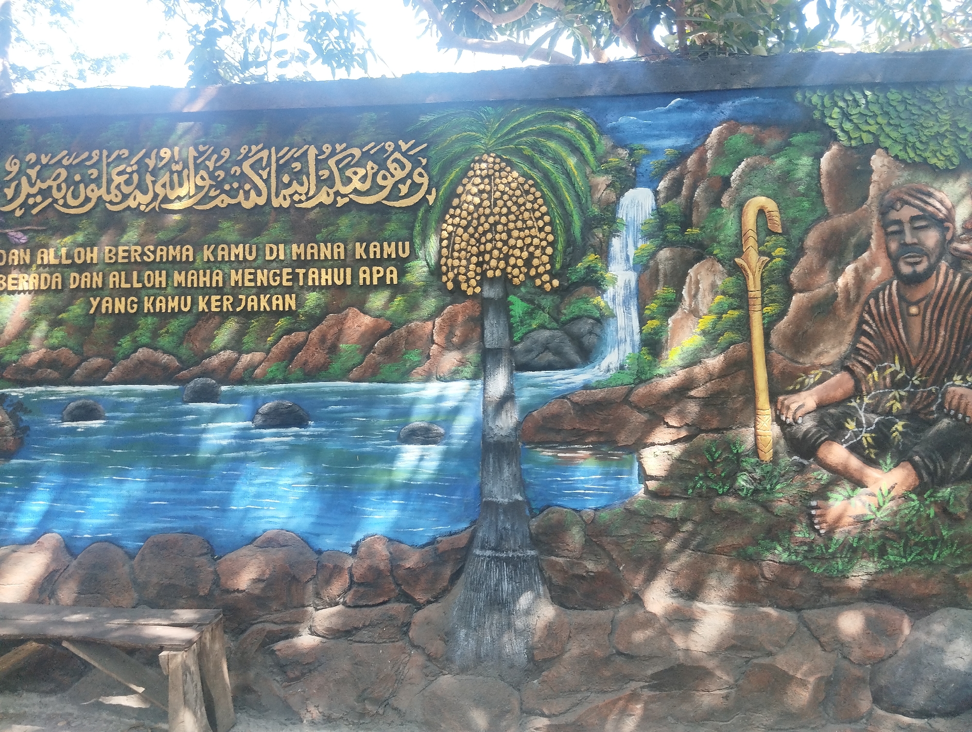 The image displays a mural on a wall in Malang, Indonesia, featuring Arabic calligraphy at the top, followed by text in Indonesian. The mural depicts a natural landscape with a river, rocks, a waterfall, and lush greenery. A palm tree laden with fruits stands prominently in the center. To the right, a man with a beard and traditional Indonesian attire, including a batik-patterned shirt and a headwrap, is seated on the ground, holding a wooden staff. Sunlight casts shadows across the mural, suggesting an outdoor setting. In the foreground, a wooden bench is visible. The overall scene evokes a tranquil and spiritual ambiance.