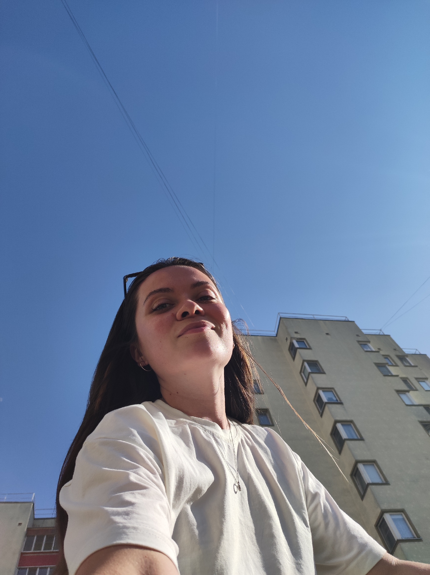 A low-angle selfie shows a young woman with long, dark hair and fair skin. She wears a white t-shirt and a silver necklace. Sunglasses are perched atop her head. The background features a clear blue sky with power lines and a tall, multi-story building with numerous windows. The building appears to be constructed in a modern or Soviet-era architectural style. Based on the architectural style of the building in the photograph, the image was probably taken in the city of Chaikovsky, Russia.