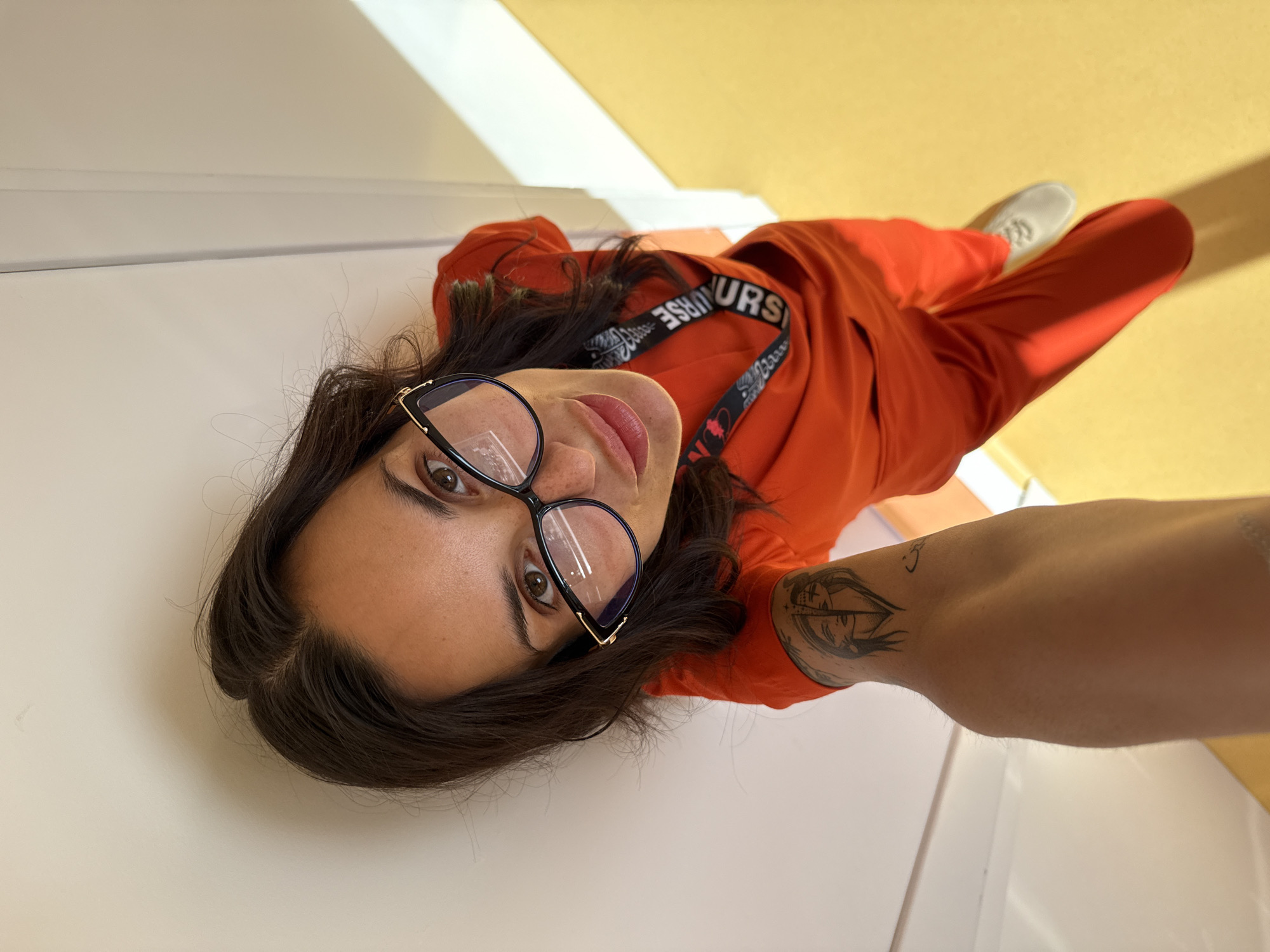 A woman takes a selfie. She is wearing black-rimmed glasses and an orange outfit with a lanyard around her neck that reads "NURSE" in black text on white material. She has dark hair and a tattoo on her left arm. The background consists of a light yellow floor and a white wall. It is not possible to determine the exact location in Pleven, Bulgaria from the image provided.