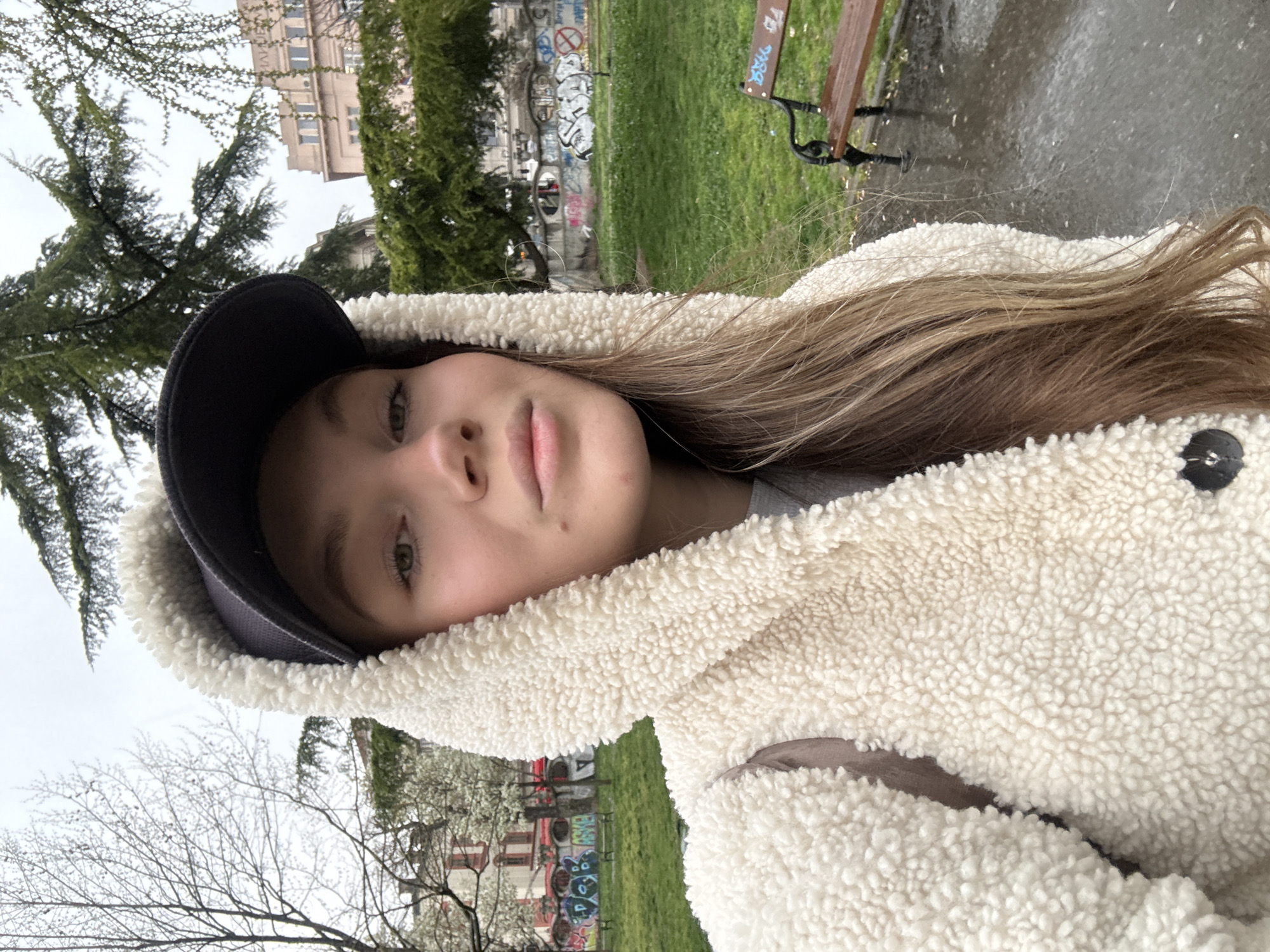 A young Caucasian female is depicted in a waist-up outdoor portrait. She is wearing a cream-colored, textured sherpa fleece hooded jacket over a gray garment, topped with a dark baseball cap. Her long, light-brown hair is visible beneath the hoodie. Her gaze is directed towards the camera with a neutral expression. The immediate foreground on the right features a wet, dark paved surface and the end of a wet wooden park bench with black metal supports. The background includes a grassy area, a low curved concrete wall extensively covered in multi-colored graffiti, and various trees, some bare with small white blossoms, and others evergreen. Behind the vegetation, multiple buildings are visible, including a light-colored, multi-story structure in the upper right background displaying the Cyrillic text "БАНЧИЋ" on its facade. The sky is overcast, and the wet ground indicates recent precipitation. The scene is located in Studentski Park (Student's Park) in the Stari Grad municipality of Belgrade, Serbia.