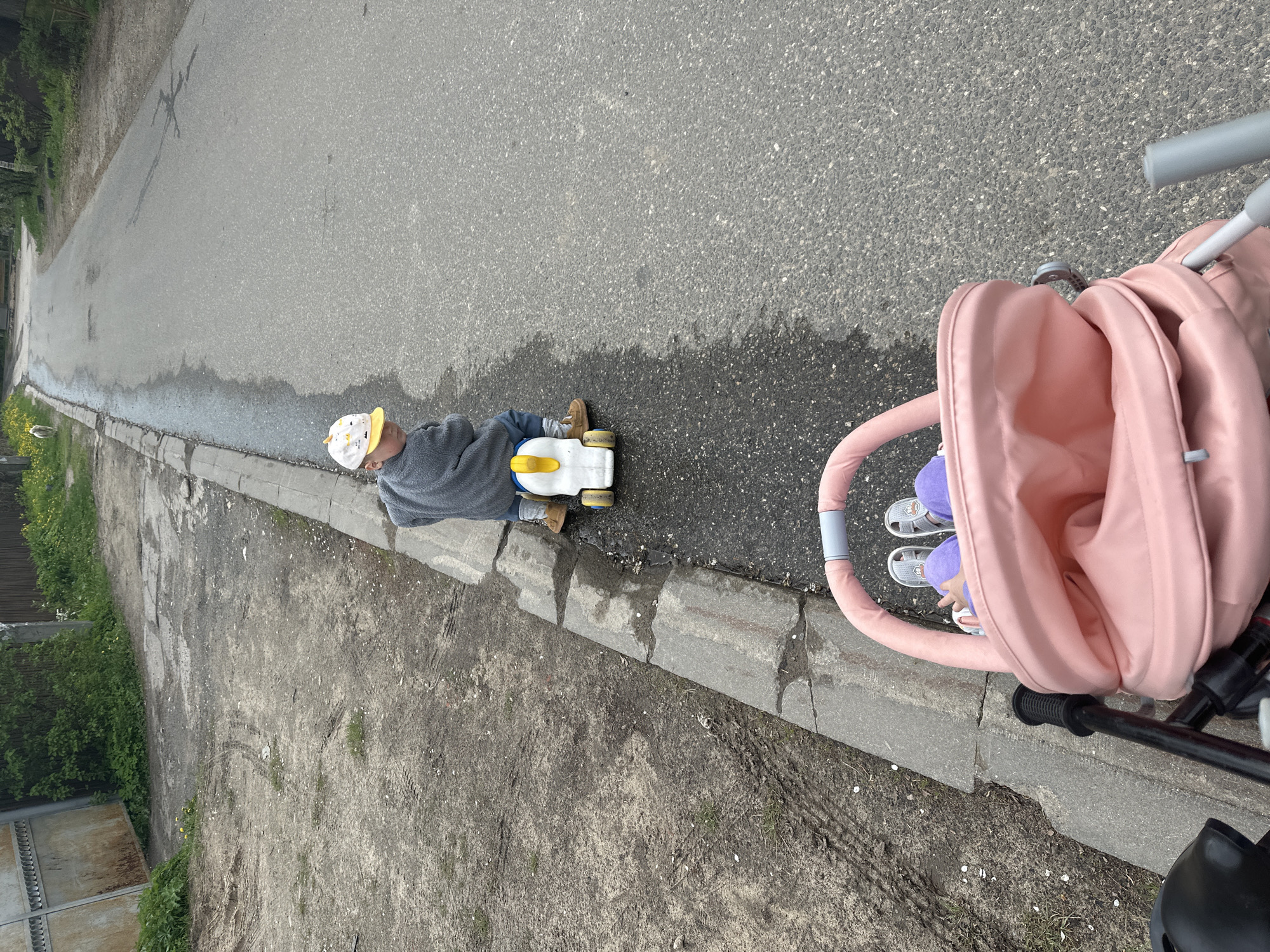 A child wearing a gray sweater, jeans, and a patterned hat is sitting on a ride-on toy vehicle on an asphalt road, adjacent to a raised concrete curb. A pink stroller with a child inside is positioned alongside the road, next to the curb. The road surface appears to be wet in places. The location is in Nizhny Novgorod, Russia. A dirt and grass area is visible next to the curb, bordered by a fence.