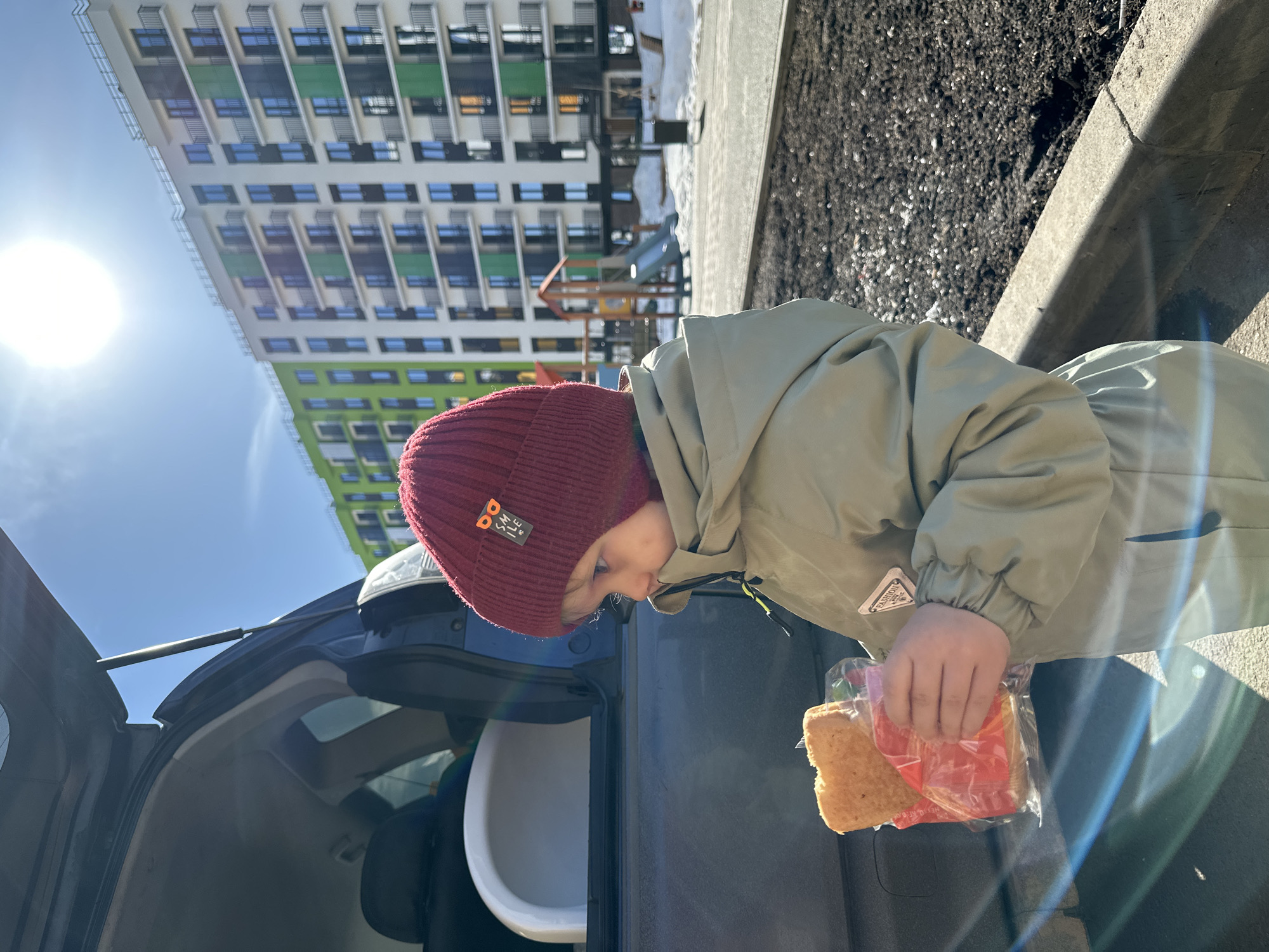 A young child is depicted outdoors in Pervomaysky, Russia, on a bright, sunny day. The child, wearing a maroon knitted beanie with a "SMILE" tag and a light olive-colored winter jacket with a "PRADA FASHION" patch, holds a packaged snack (a rectangular pastry in a clear plastic bag with red interior packaging) in both hands. The child is positioned next to the rear of a dark-colored vehicle, possibly an SUV or hatchback, with its tailgate open. A light-colored child's car seat or booster seat is visible within the vehicle's cargo area. In the background, modern multi-story apartment buildings with white, green, and dark facades and numerous balconies are visible under a clear blue sky. Patches of snow are present on the ground, alongside bare dark earth and an asphalt paved area. A children's playground structure with a slide is also visible in the mid-ground. The sun is high, casting a prominent flare in the upper right portion of the image.