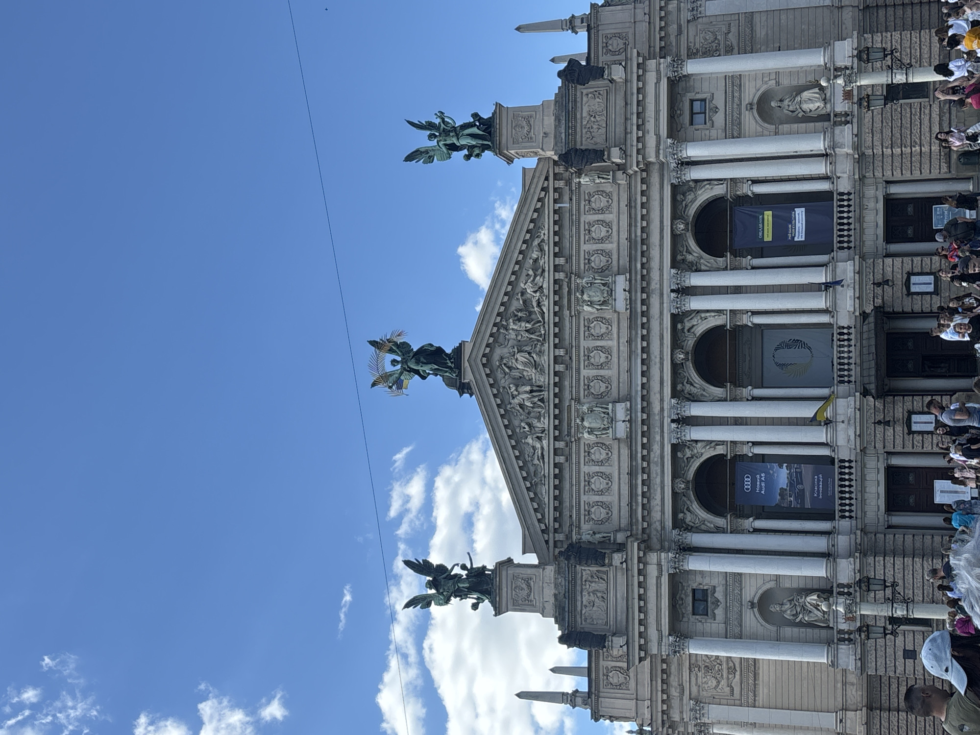 The image shows a section of the Lviv Opera House in Ukraine. The photograph is taken from a slightly low angle, emphasizing the ornate facade of the building. Three statues of angels adorn the roofline, set against a background of blue sky with scattered clouds. Banners are suspended from the columns of the building. Crowds of people can be seen on the right side of the frame, suggesting an event or gathering. The architecture is classical in style, with detailed carvings and sculptures embellishing the structure.