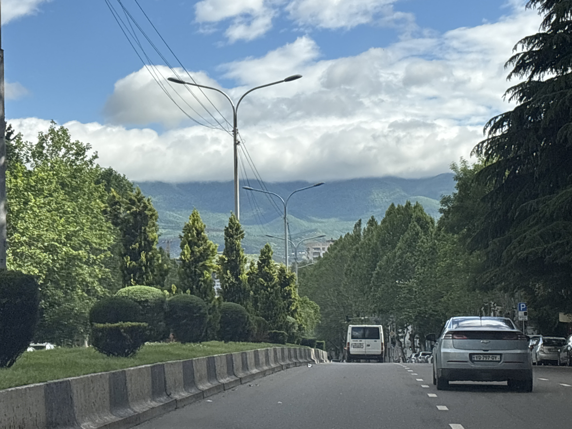 The image depicts a street scene in Tbilisi, Georgia, characterized by a wide road with several lanes. The road is lined with trees and landscaped green spaces separated by a concrete median barrier painted with black and white stripes. Overhead, there are multiple streetlights and power lines. In the distance, there are mountain ranges partially obscured by clouds. The sky is blue with scattered white clouds.
Vehicles are present on the road, including a light grey Chevrolet Volt with the license plate "YQ-797-QY," which is partially visible. Further down the road, a white van and other cars are visible. There are no visible people in the immediate foreground. A "P" parking sign is visible on the right side of the road near some parked vehicles.