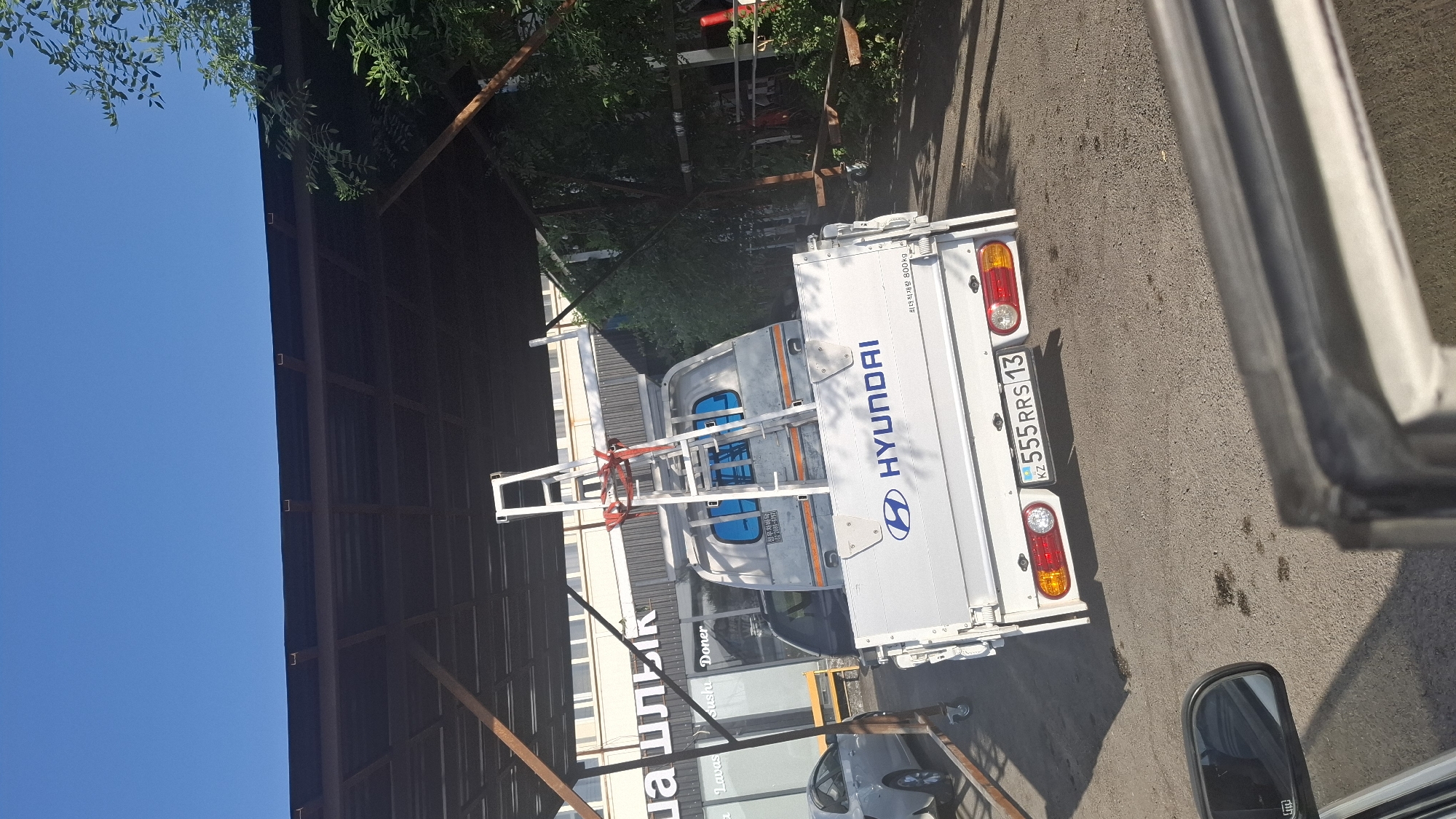 A white Hyundai truck is parked under a dark brown metal awning, with a ladder secured on top. The truck has a license plate that reads "KZ 555 RRS 13". In the background, a building with white windows and signage is visible, with some trees to the right of the frame. The ground is asphalt. The scene appears to be a sunny day.