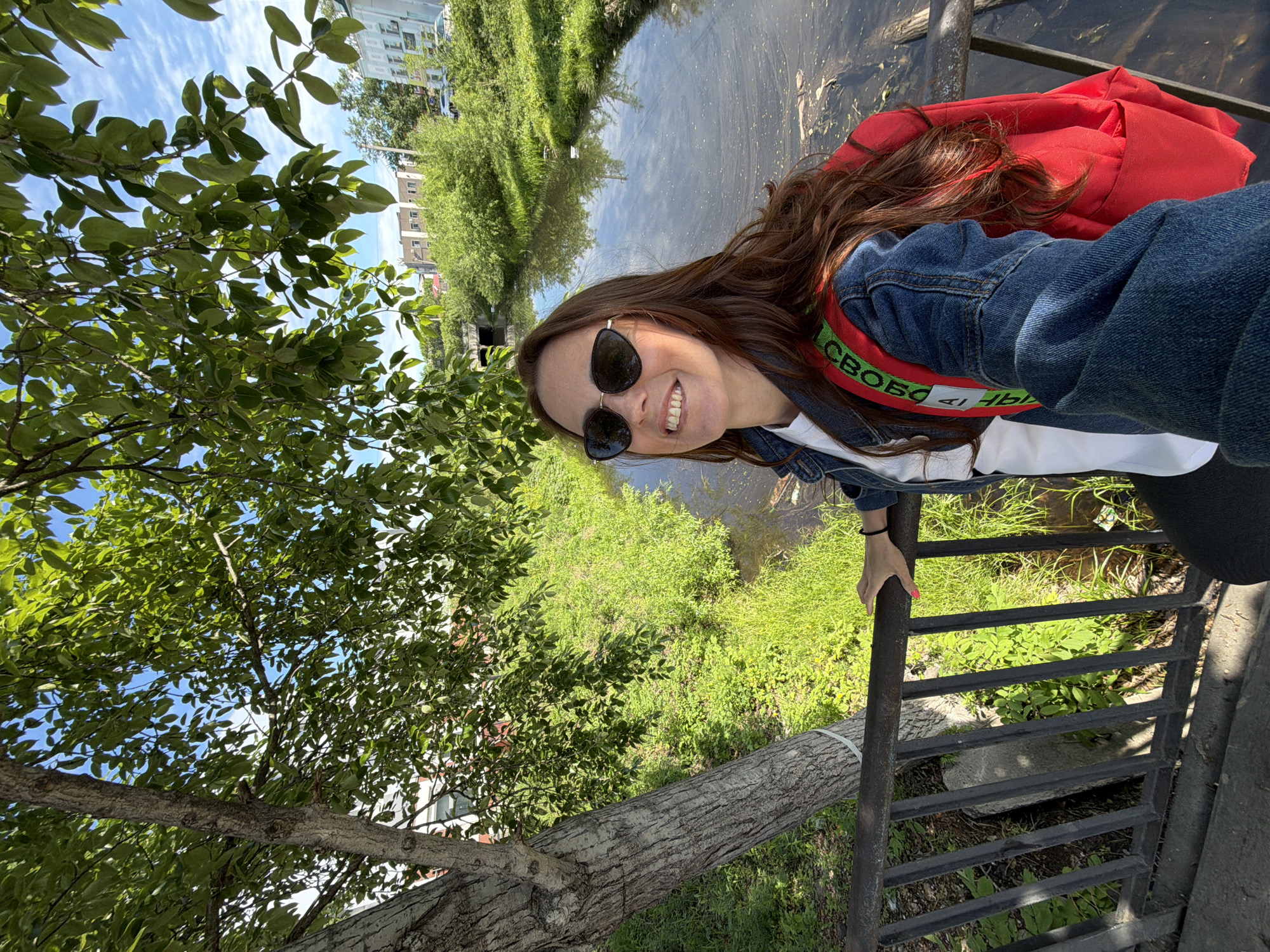 In Birobidzhan, Russia, a woman with long, brown hair and sunglasses is taking a selfie on a bridge over a small canal. She is wearing a denim jacket over a white shirt and a bright red backpack with green straps. The canal is surrounded by lush green vegetation, and a tree with textured bark stands to her left. In the background, buildings and more greenery are visible, suggesting an urban or semi-urban setting. The sky is blue with scattered clouds.